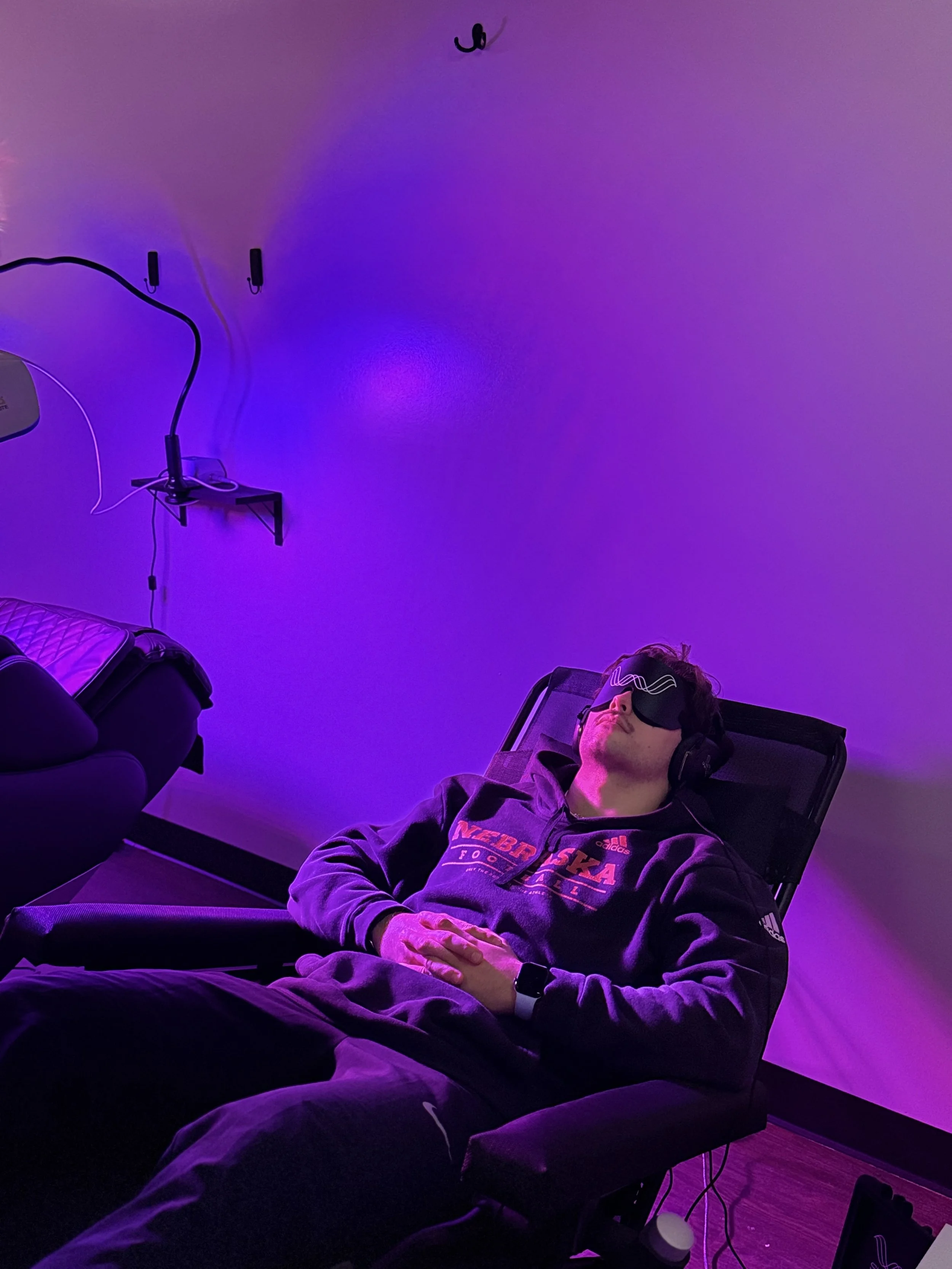 A person with closed eyes lying on a reclined chair, wearing a hospital gown and a sleep mask, with purple ambient lighting.
