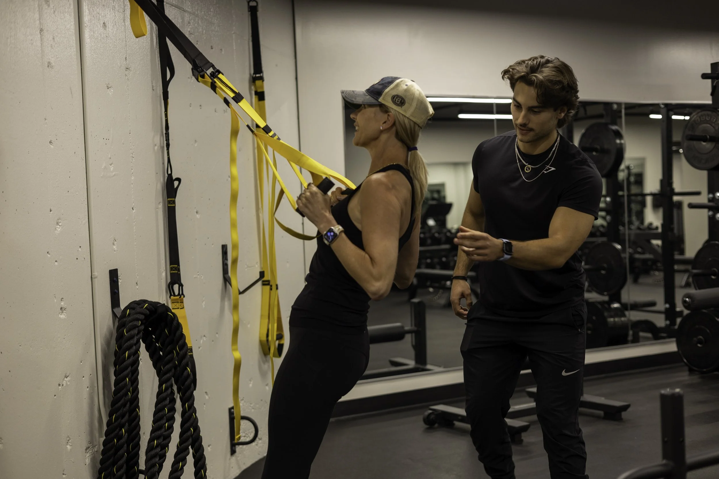 A woman doing suspension training using yellow and black straps attached to a wall in a gym while a male trainer assists her.