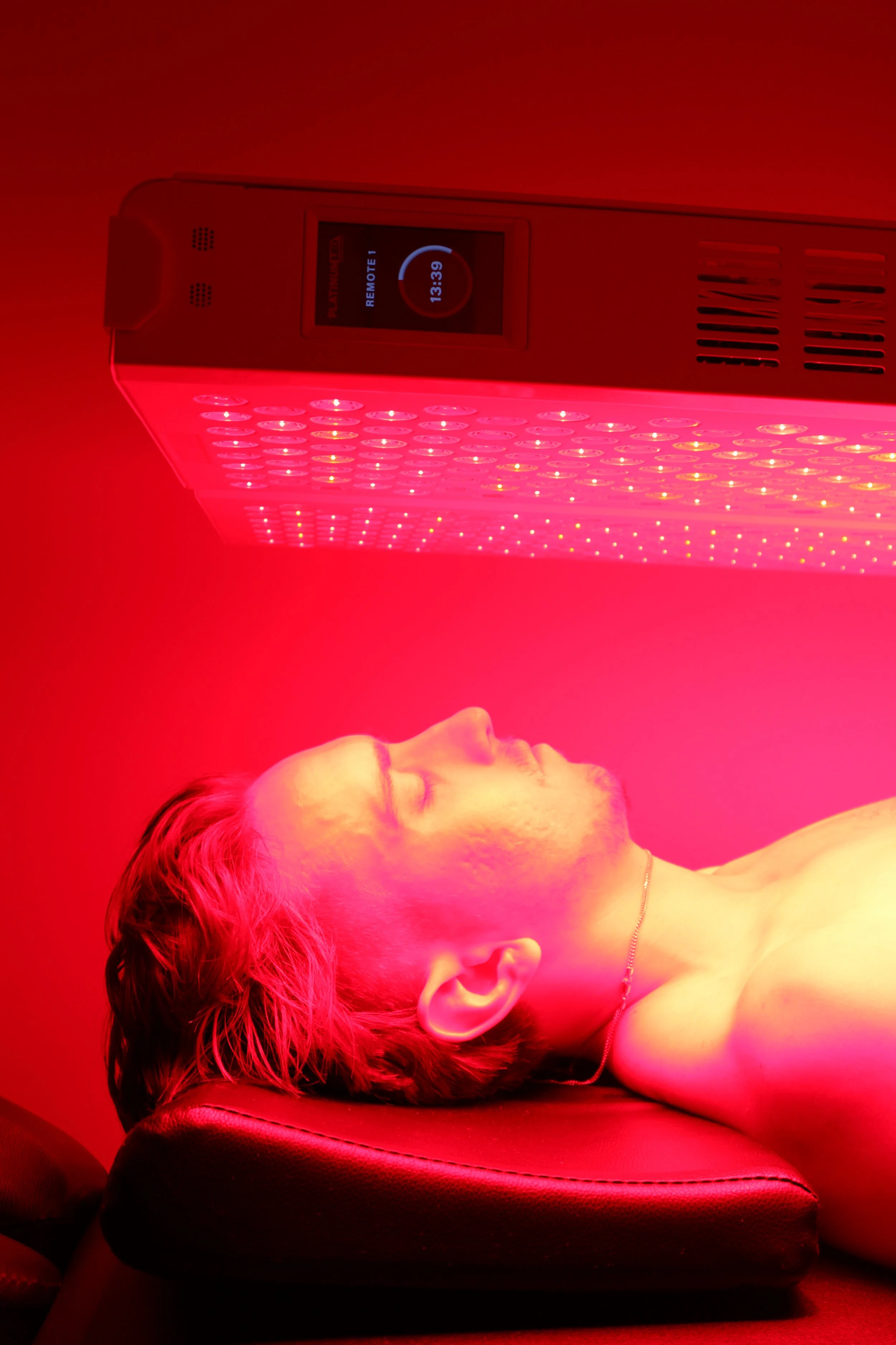 A person with red hair lying down under a red LED light therapy panel in a dark room.