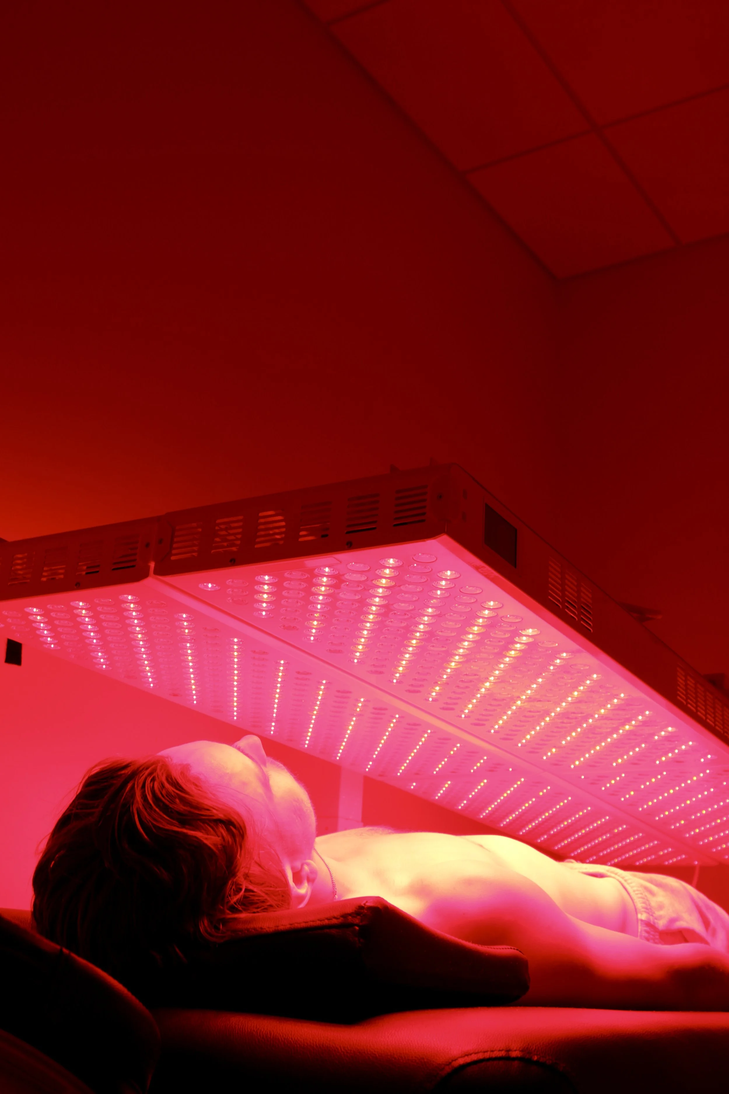Person lying on a bed under red LED light, with a red-tinted ceiling with square panels visible.
