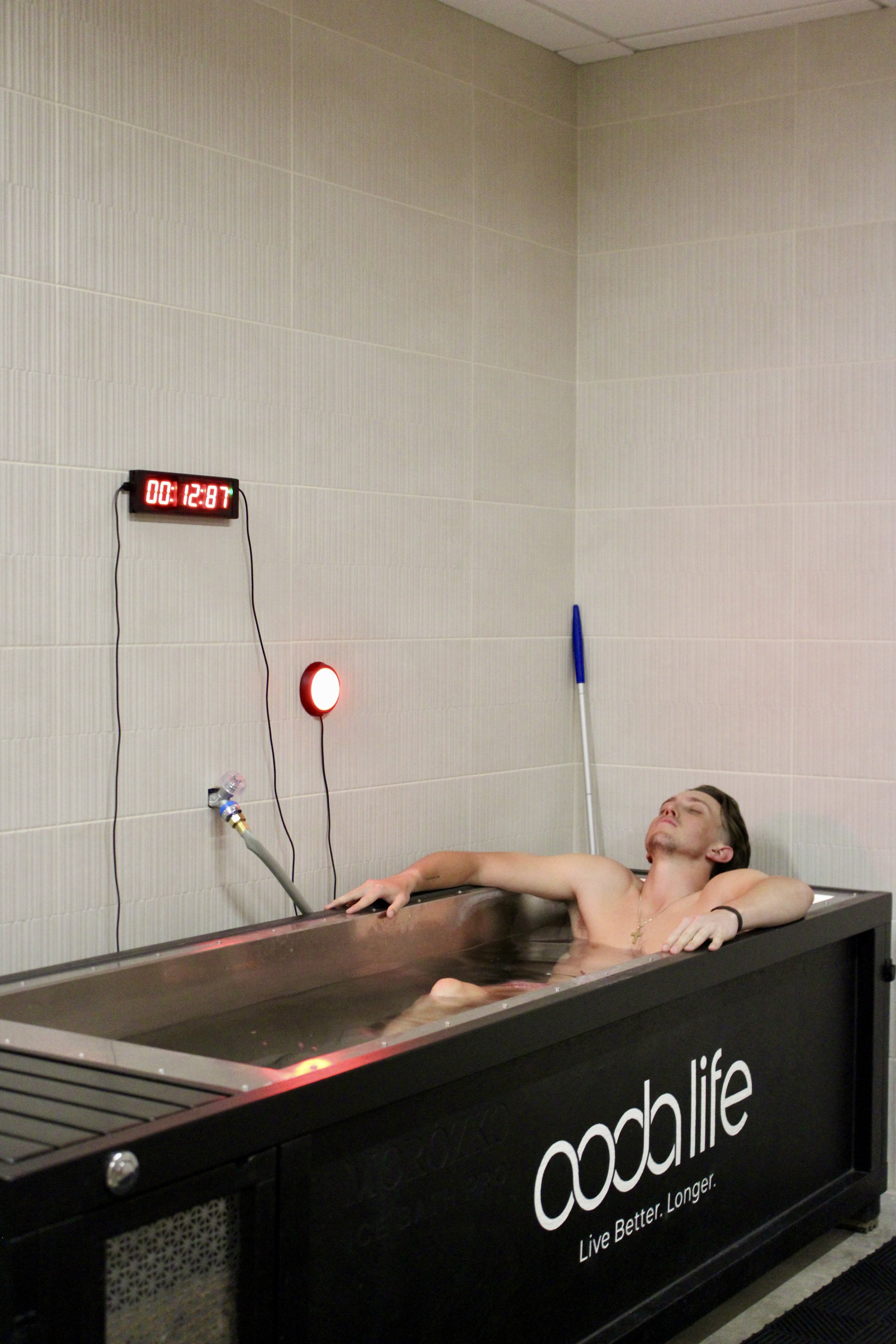 A man lying in a cold plunge tub with his eyes closed, wearing a cross necklace, in a room with beige tiled walls. A digital timer on the wall shows 12:87. The tub has black sides with white text that reads 'ooblife' and 'Live Better. Longer.'