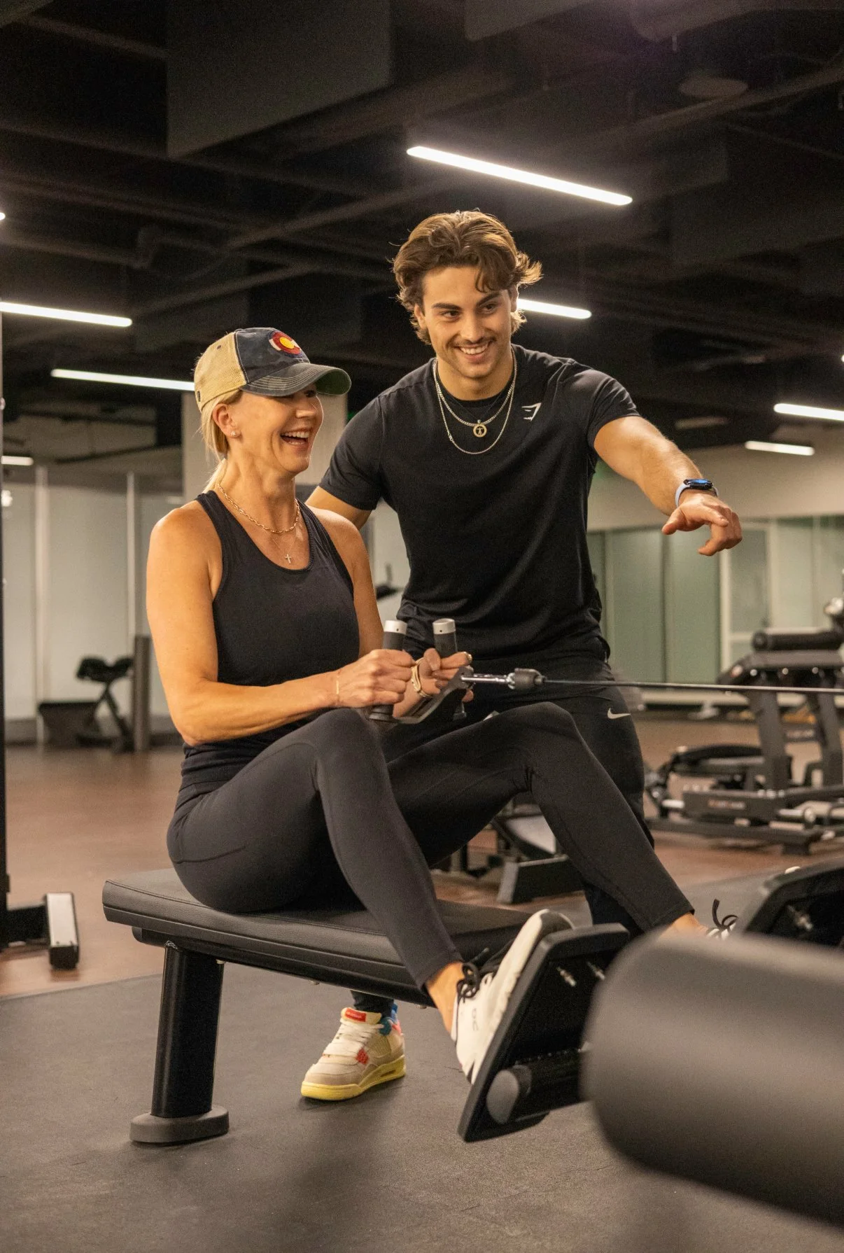 A woman sitting on a rowing machine in a gym, smiling, with a young man standing next to her, pointing, both appearing happy and engaged in a workout session.