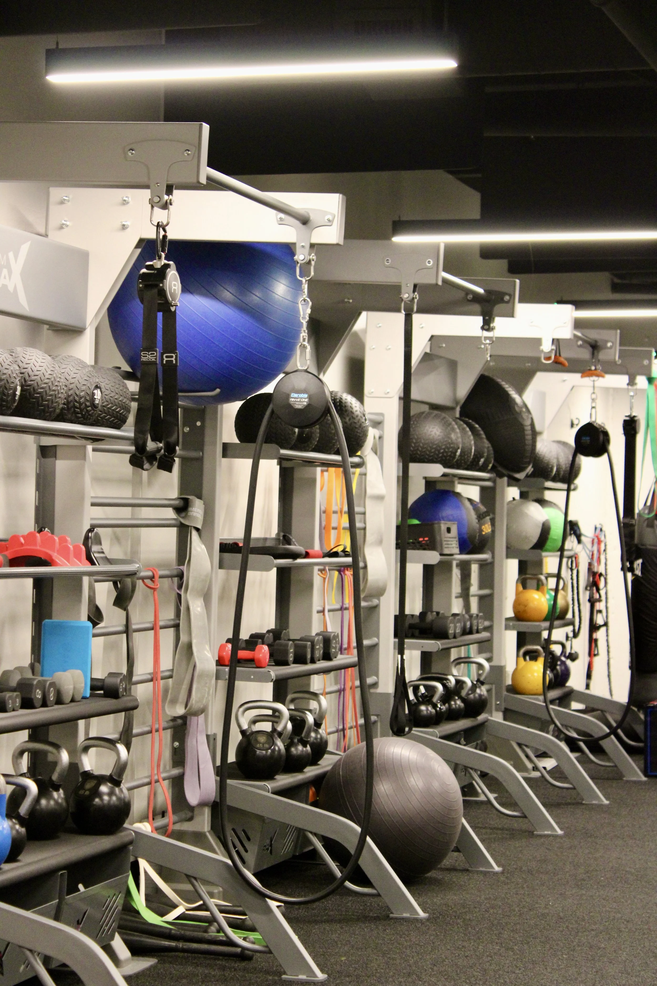 Gym equipment including kettlebells, medicine balls, dumbbells, resistance bands, and a large blue exercise ball arranged on shelves and racks in a fitness area with black flooring and overhead lighting.