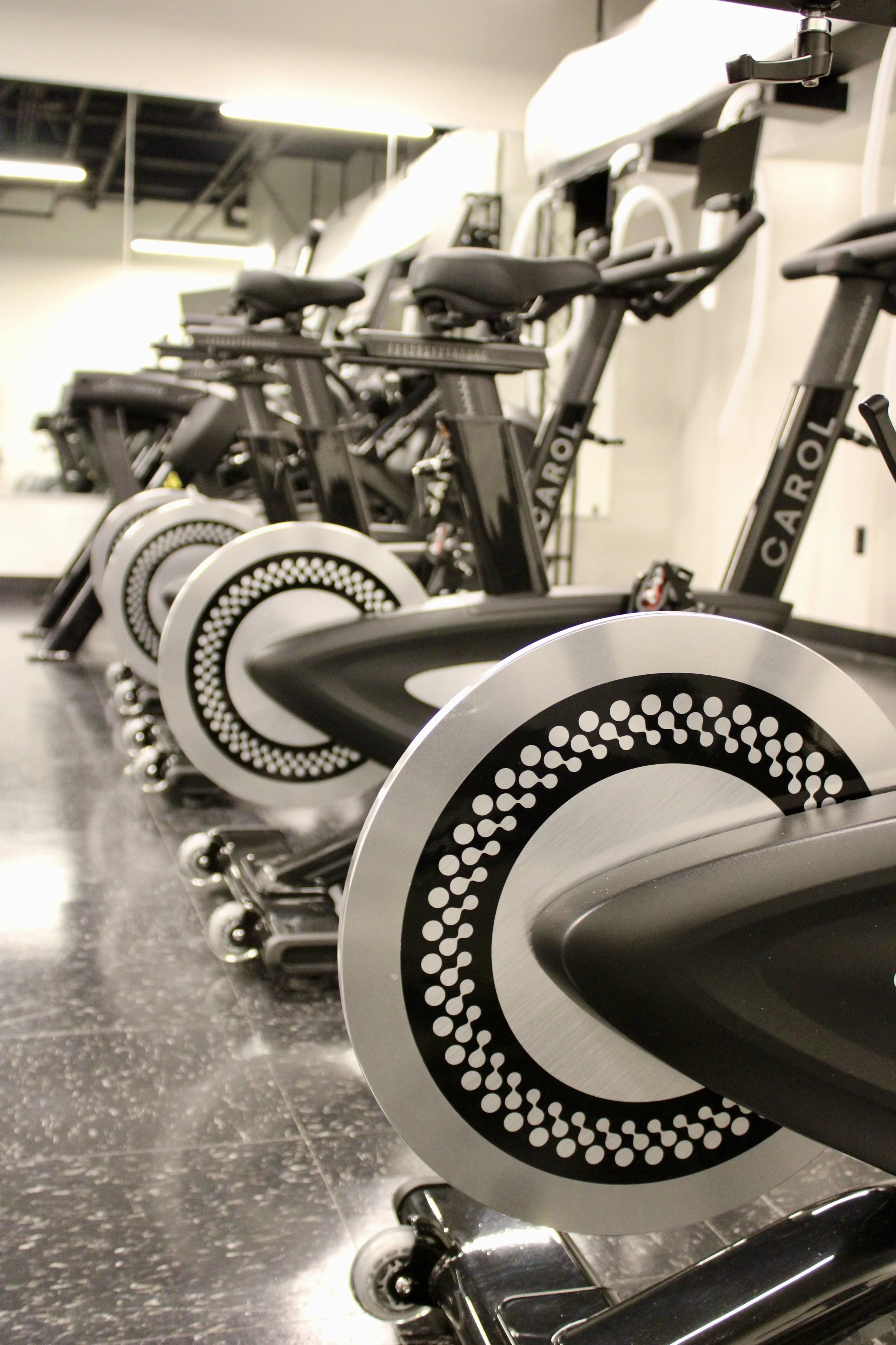 A row of multiple black stationary exercise bikes lined up, with black and white circular designs on the flywheels, in a fitness gym with bright overhead lighting and a black tiled floor.