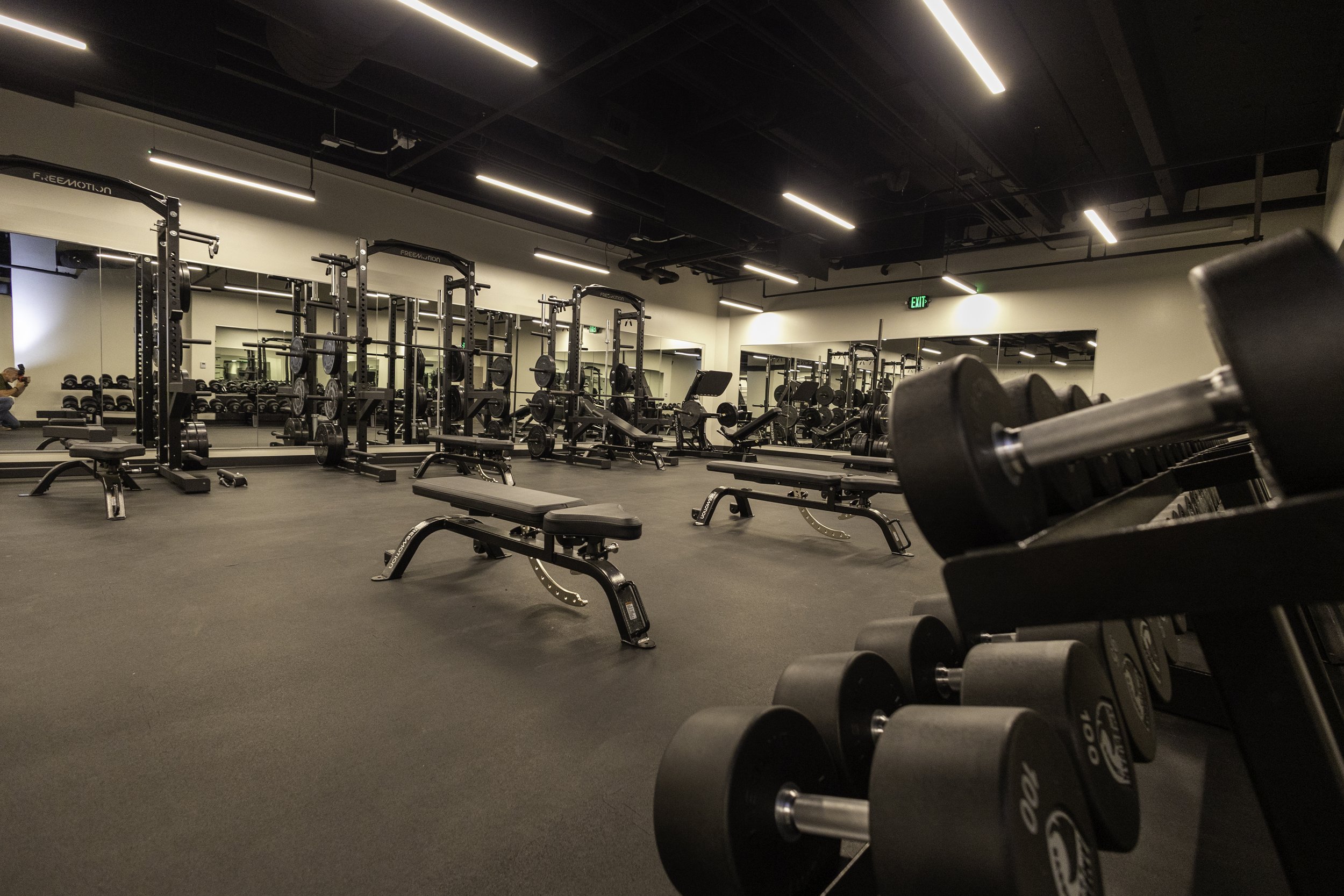 Empty gym with lifting equipment, benches, and free weights, reflected in mirrors on the wall, with a person taking a photo in the mirror on the left.