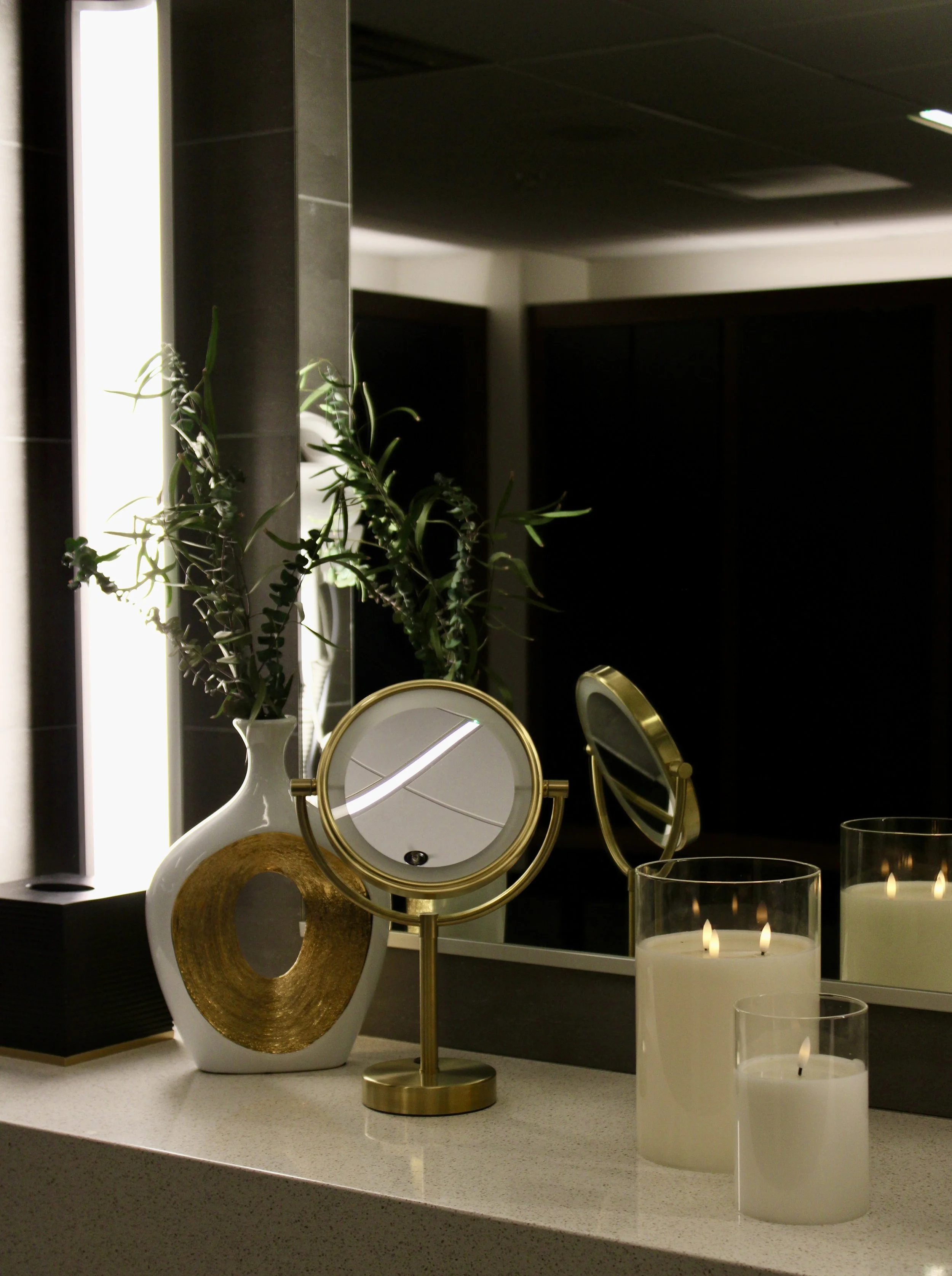 Decorative items on a kitchen countertop, including a white vase with green plant, a gold mirror, a gold candle holder, and white candles in glass holders.