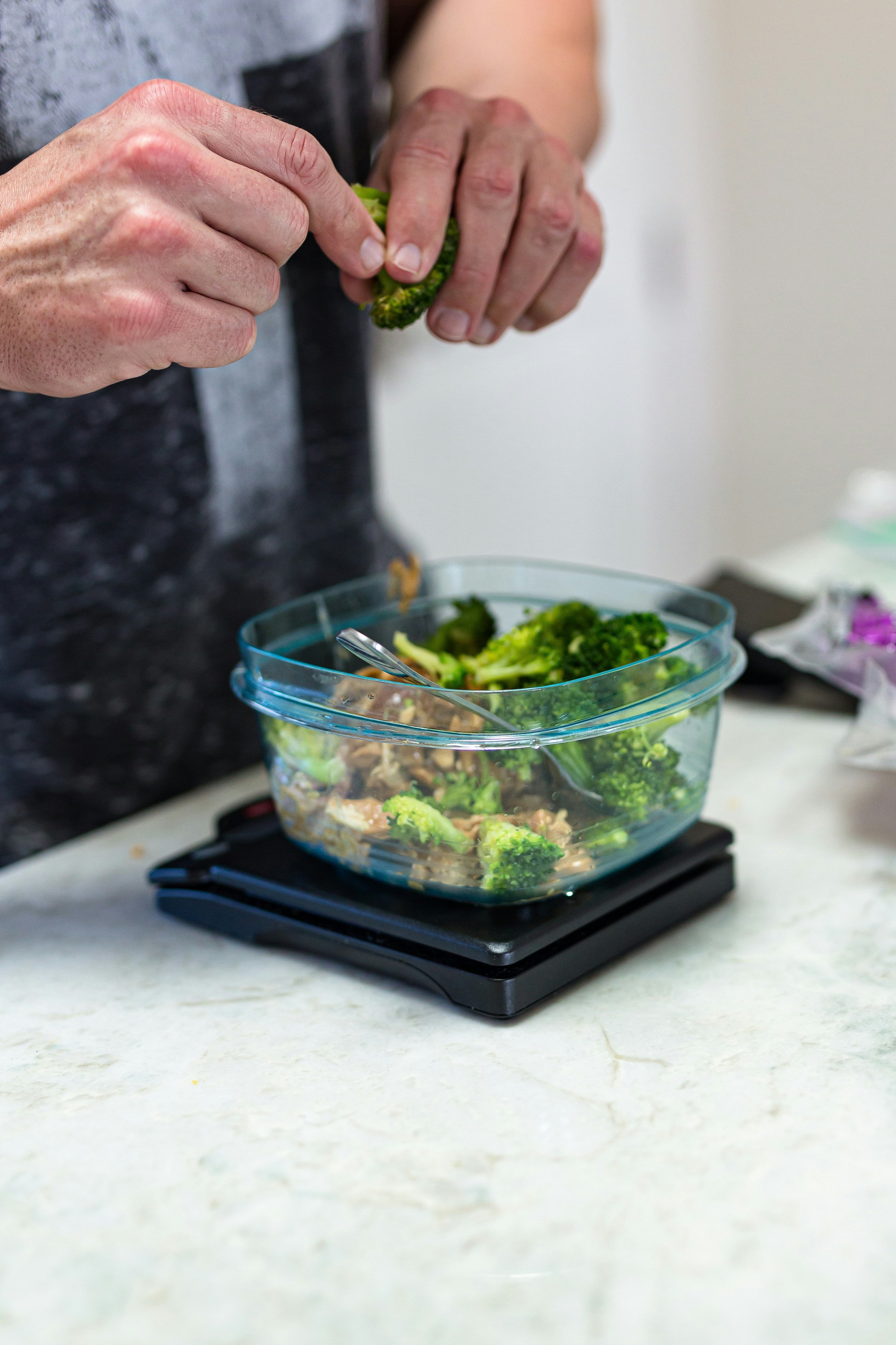 Person placing broccoli florets into a glass plastic container on a digital scale in a kitchen.