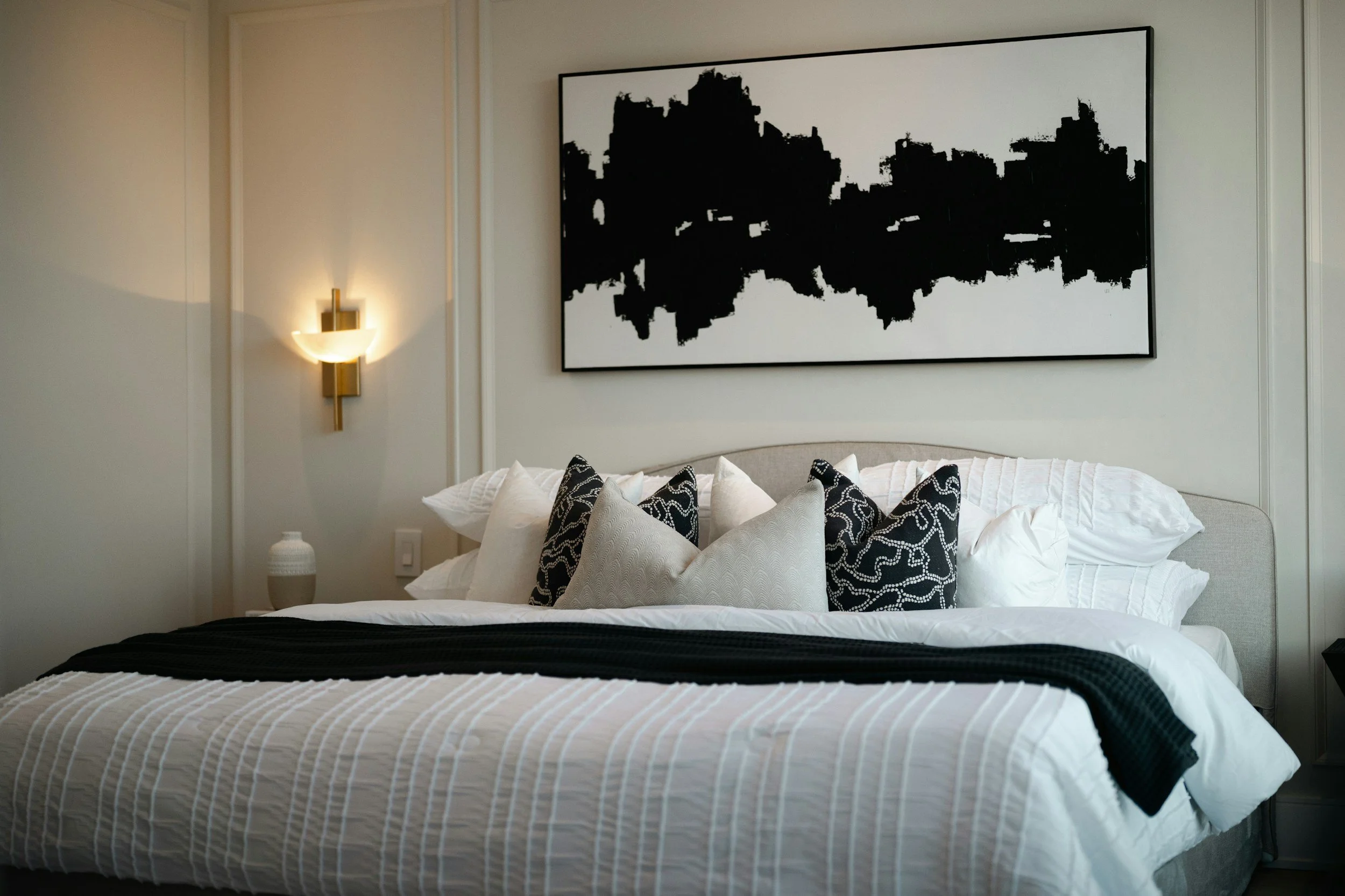 A neatly made bed with white linens and multiple pillows, including black and white patterned pillows, in a modern bedroom with a large abstract black and white framed artwork on the wall. A wall-mounted light fixture is on the left side of the artwork.