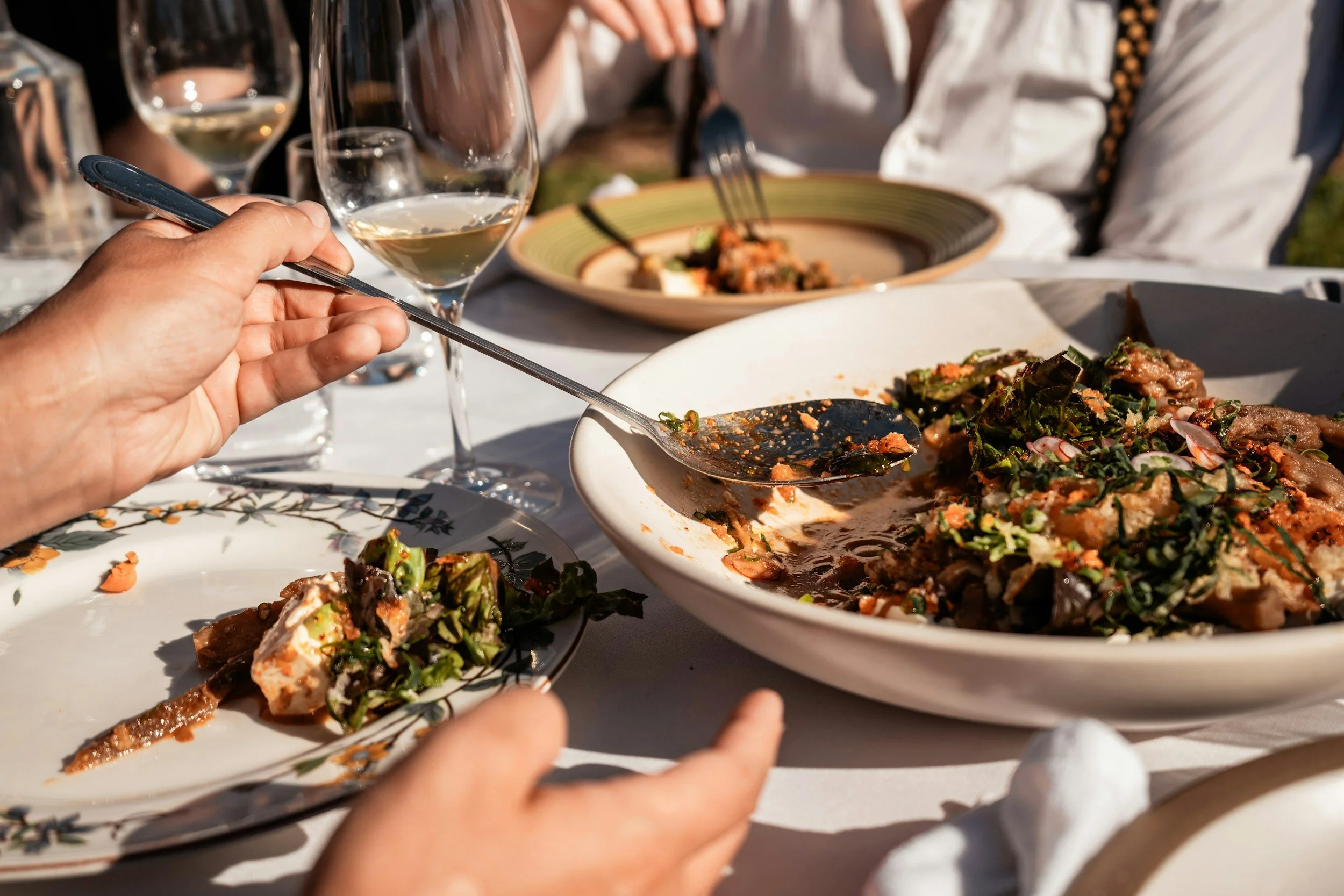 People enjoying a meal outdoors, with plates of salad and glasses of white wine on a table.