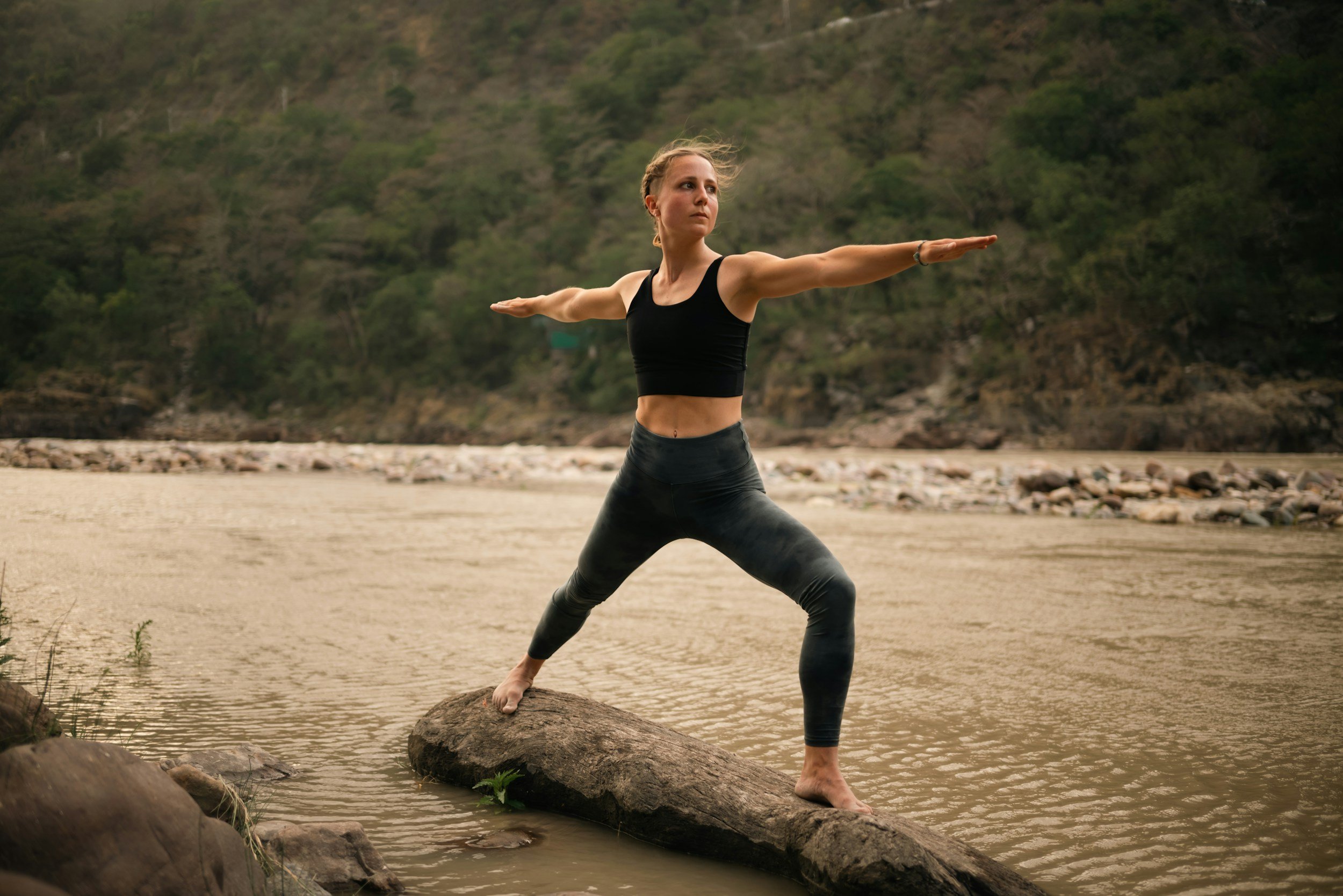 A woman practicing yoga on a rocky riverbank, standing on a large rock with one foot, with her arms extended horizontally, wearing black workout clothing, overlooking a river with hills covered in trees in the background.
