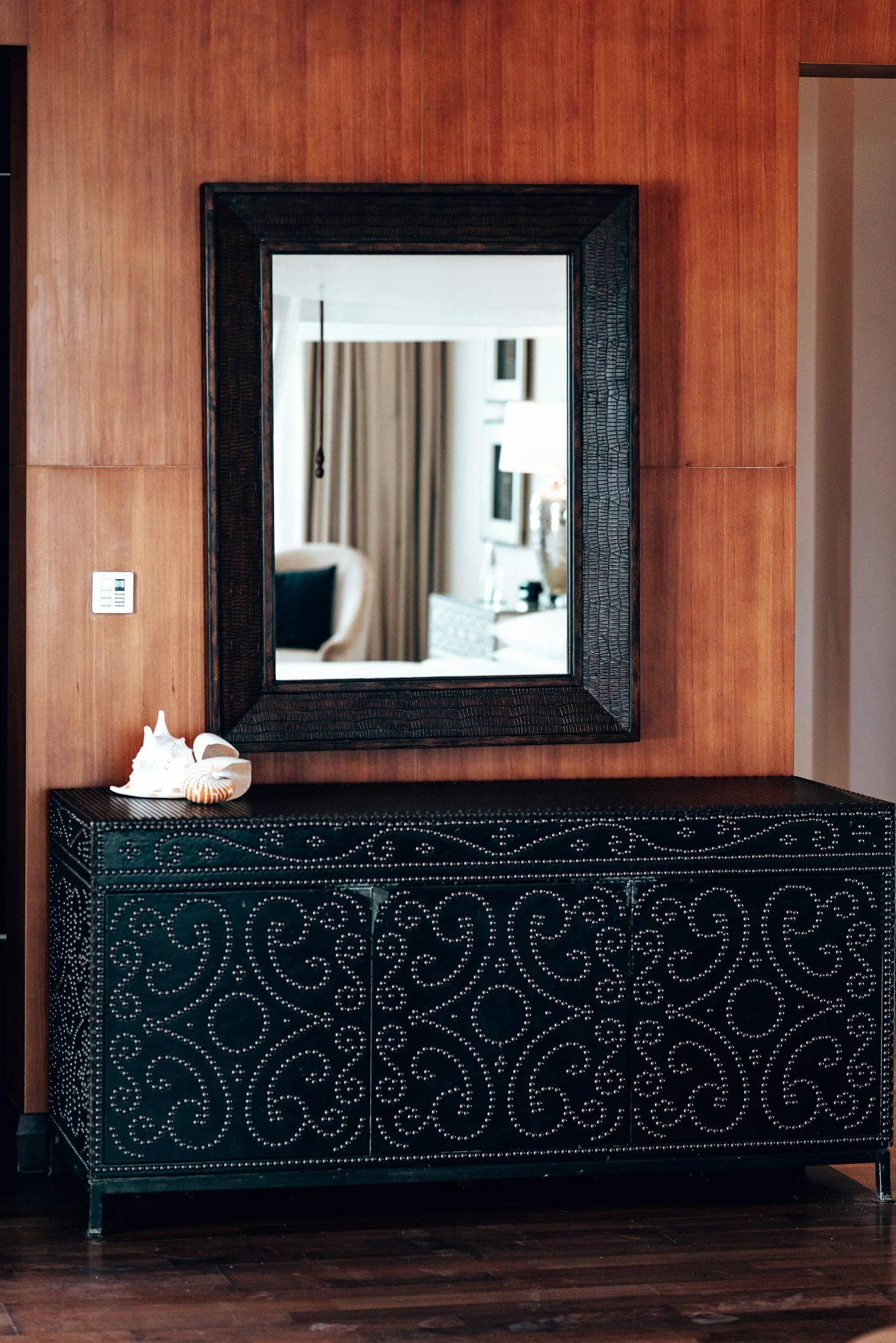 A black cabinet with intricate swirly patterns on its surface sits in front of a wooden wall. A mirror with a dark textured frame hangs on the wall above the cabinet, reflecting part of a room with a window, curtains, and a lamp. On top of the cabinet are decorative seashells.