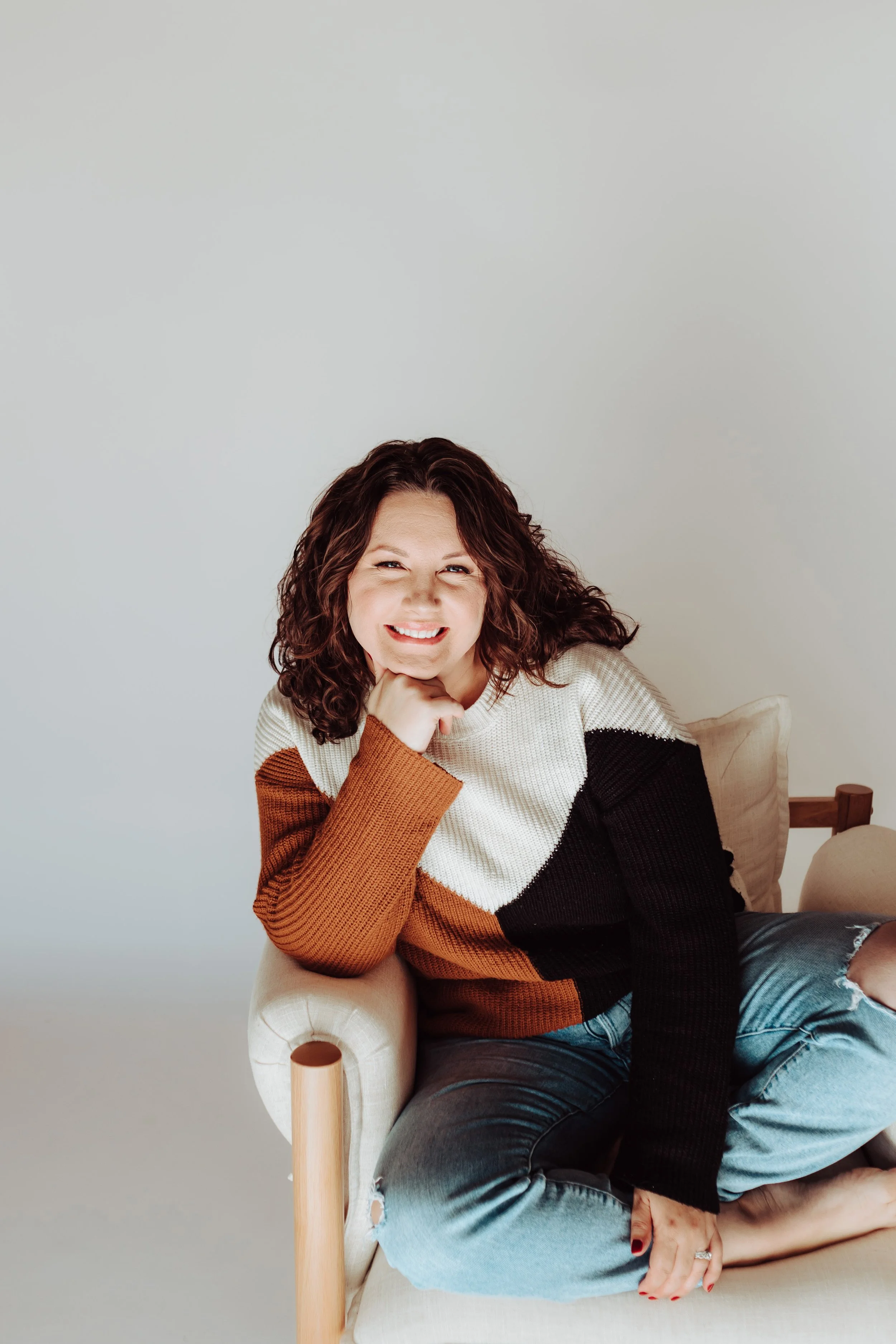 A woman with curly hair smiling, sitting in a beige chair in a room with a plain light gray wall, wearing a color-blocked sweater with brown, white, black, and orange sections, and ripped jeans.