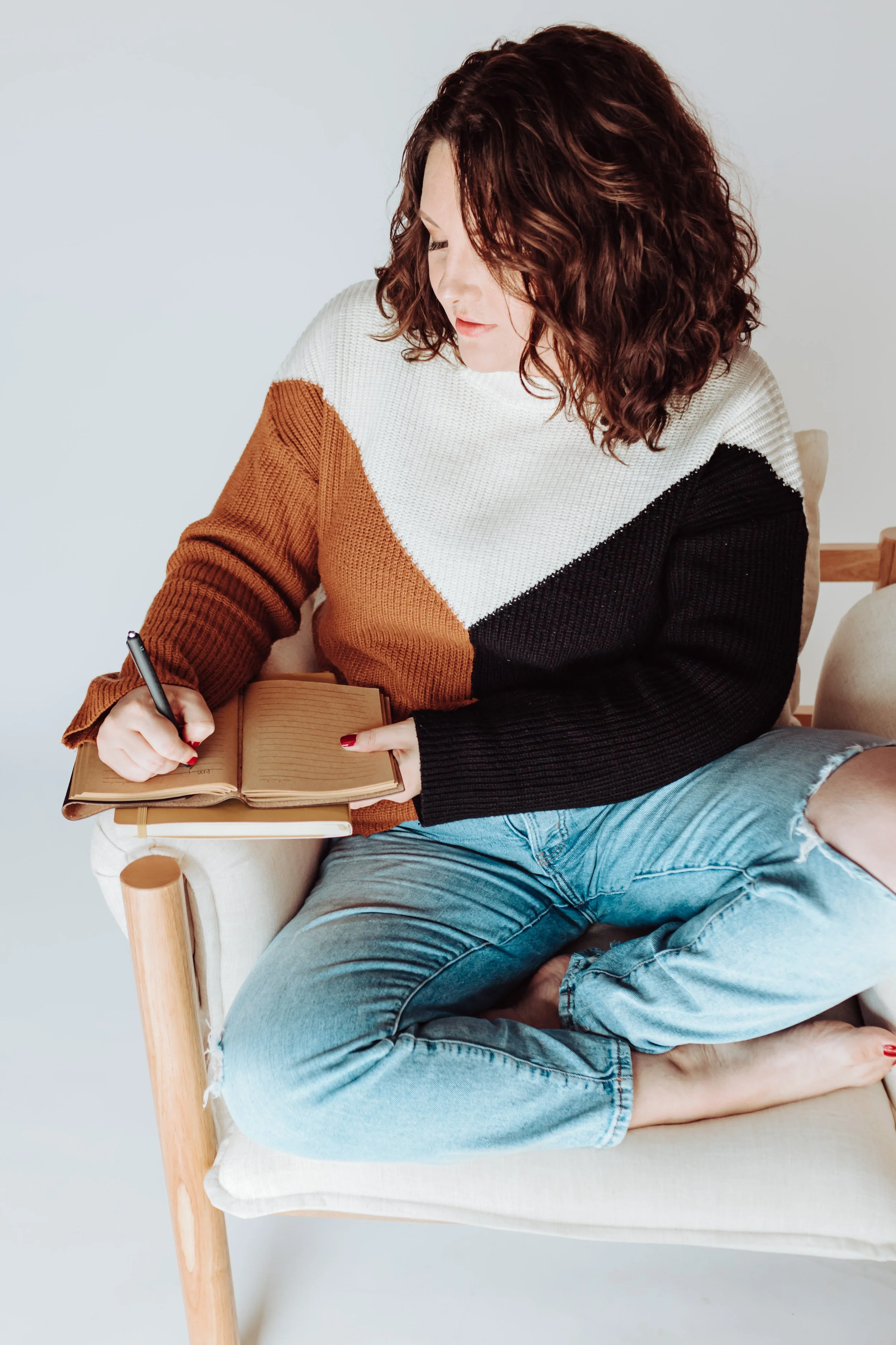 A woman with curly brown hair sitting on a beige armchair, writing in a notebook, with a person lying on her lap, wearing ripped jeans. The background is plain white.