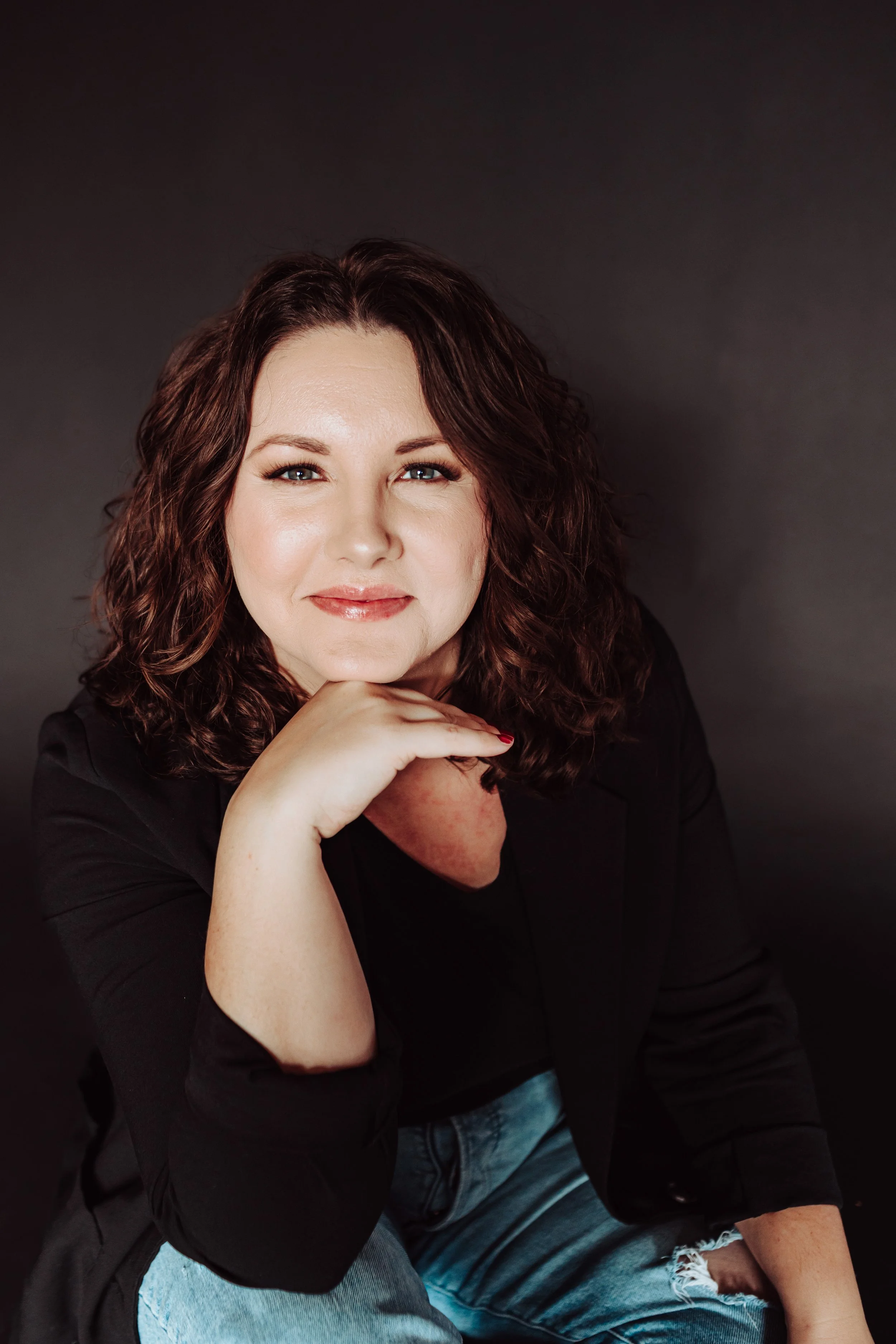 A woman with curly brown hair and fair skin, wearing a black top, sitting against a dark background, smiling, with her chin resting on her hand.