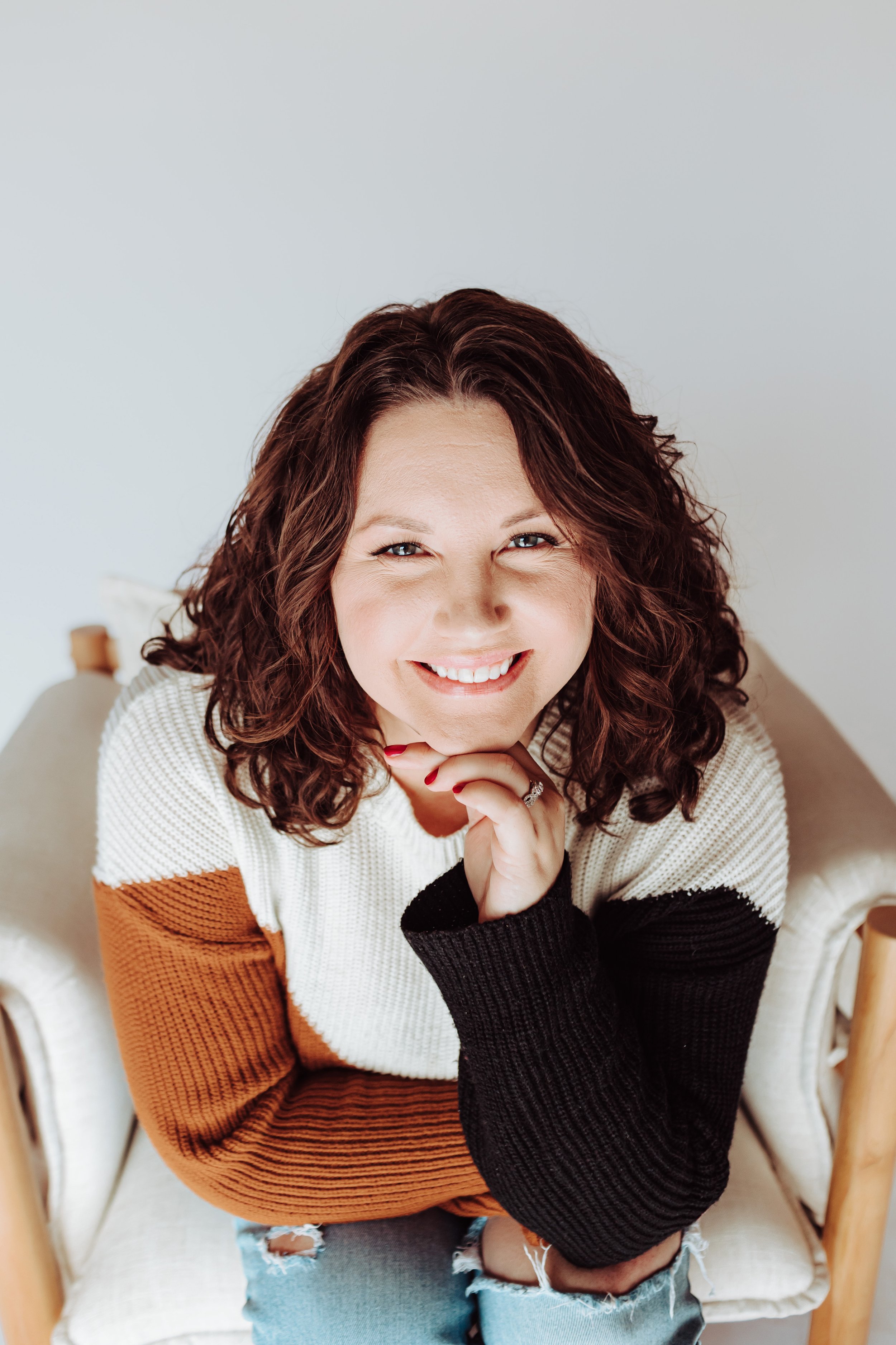A woman with curly brown hair and blue eyes smiling, sitting on a beige chair, wearing a color-blocked sweater with white, black, and rust sections, and ripped jeans.