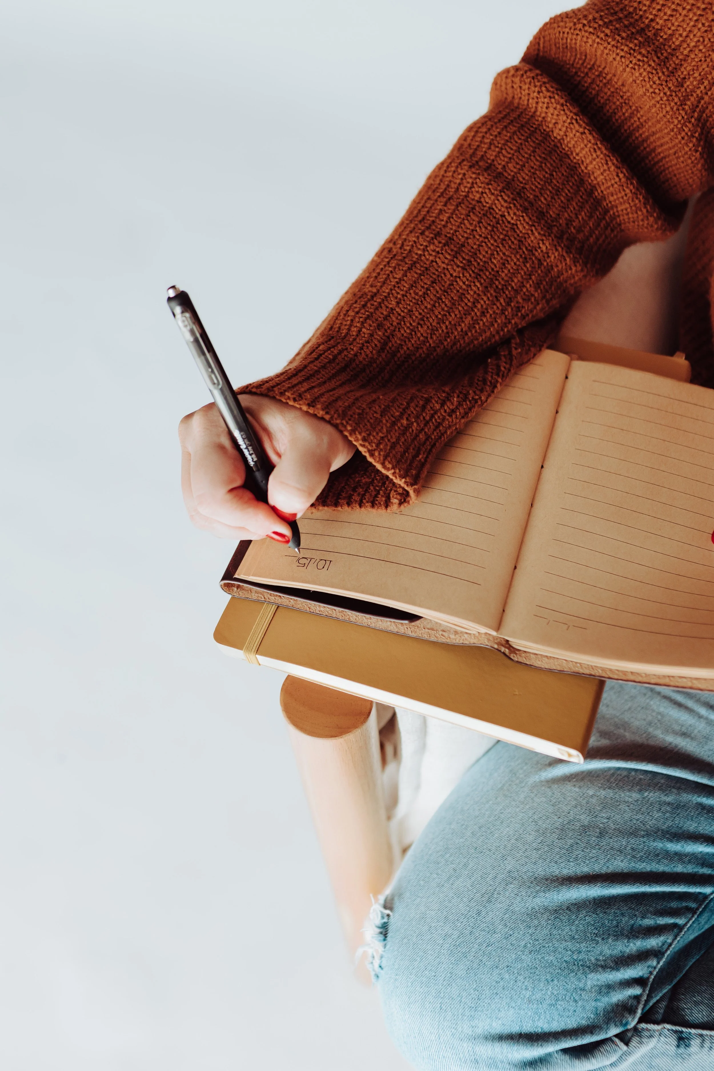 A person wearing a brown knit sweater and ripped blue jeans sitting at a small wooden table, writing in a lined notebook with a black pen.