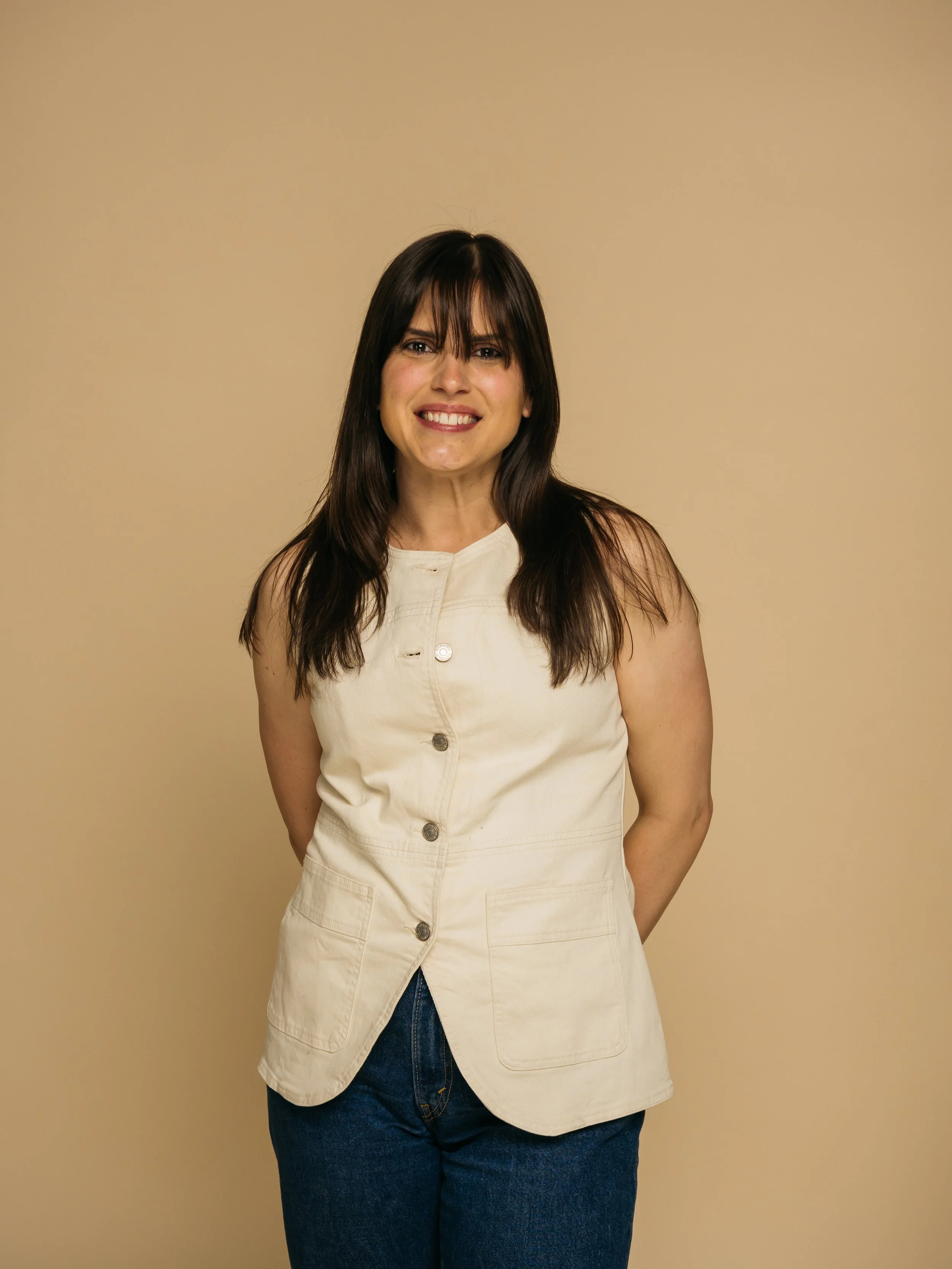 A woman with long dark hair and bangs, smiling, wearing a cream sleeveless button-up dress and dark jeans, standing against a plain beige background.