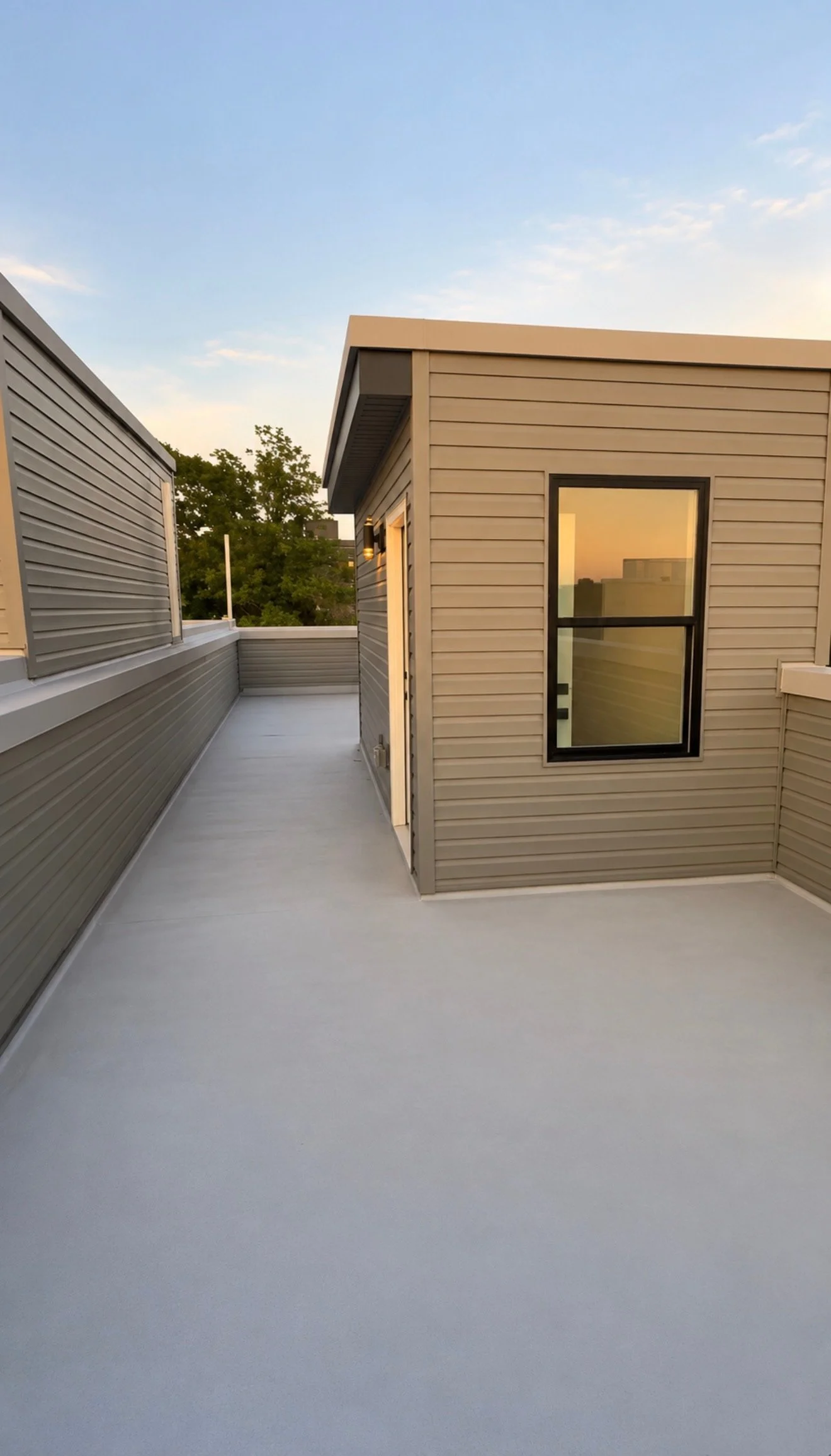 A rooftop patio with beige siding, a door, a window, and a concrete floor, with a clear sky and trees in the background.