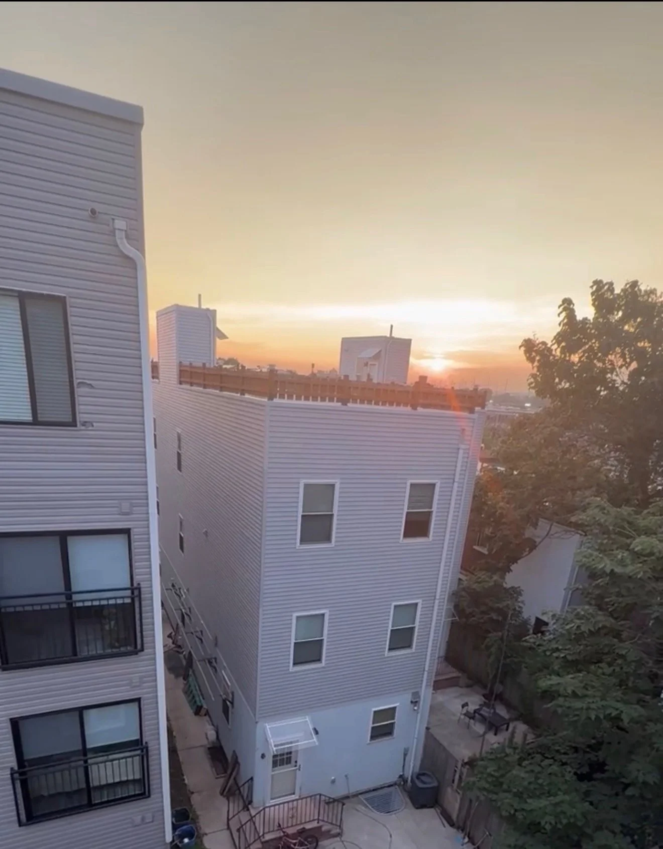 Sunset view of modern white residential buildings with multiple windows and balconies, trees, and a fence, in an urban neighborhood.