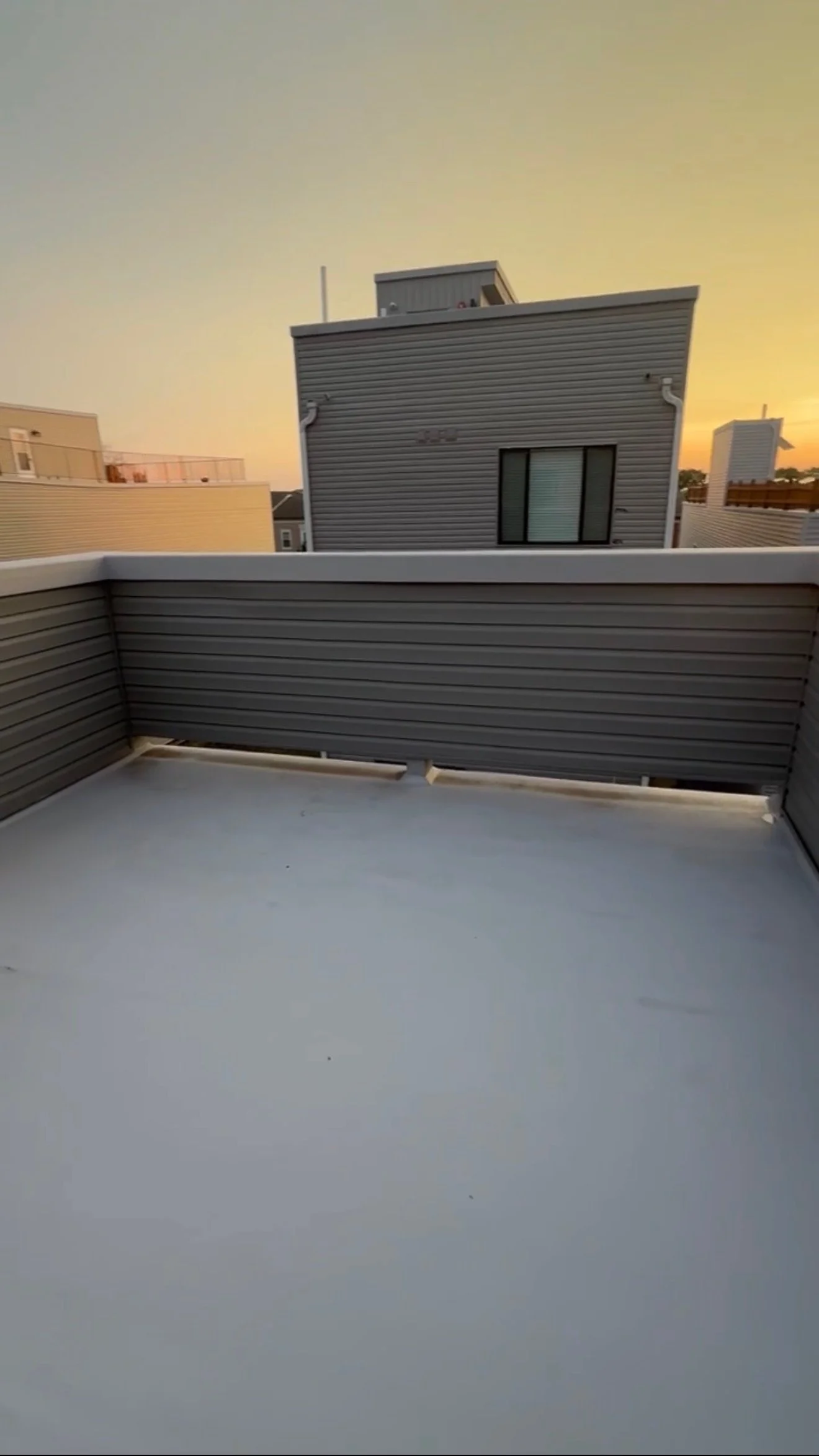 View of a small balcony with gray metal railing, smooth gray flooring, and neighboring modern buildings with metal siding during sunset.
