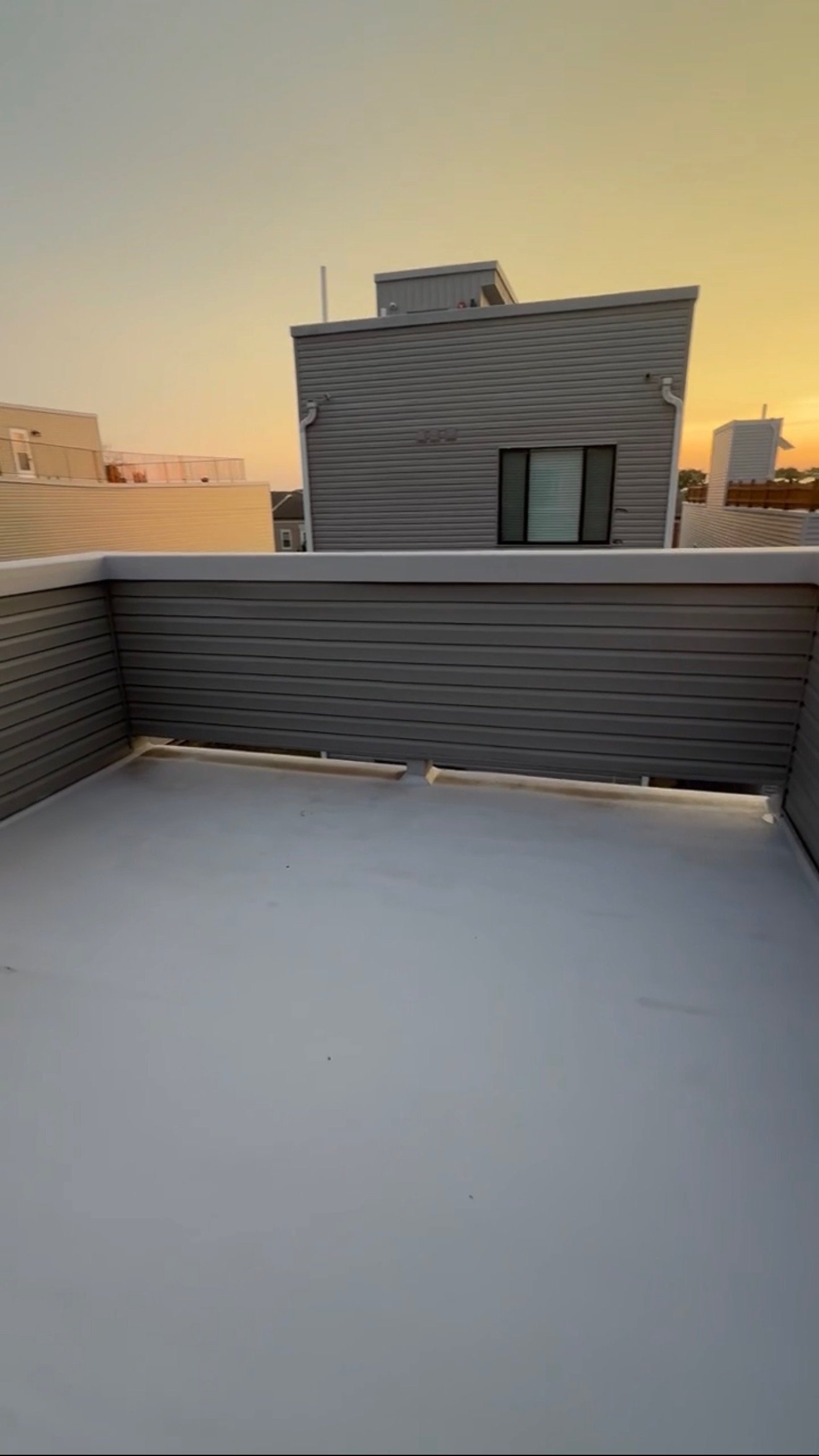 View of a rooftop balcony with taupe metal siding, a concrete floor, and a gray metal railing, with neighboring buildings and a sunset in the background.