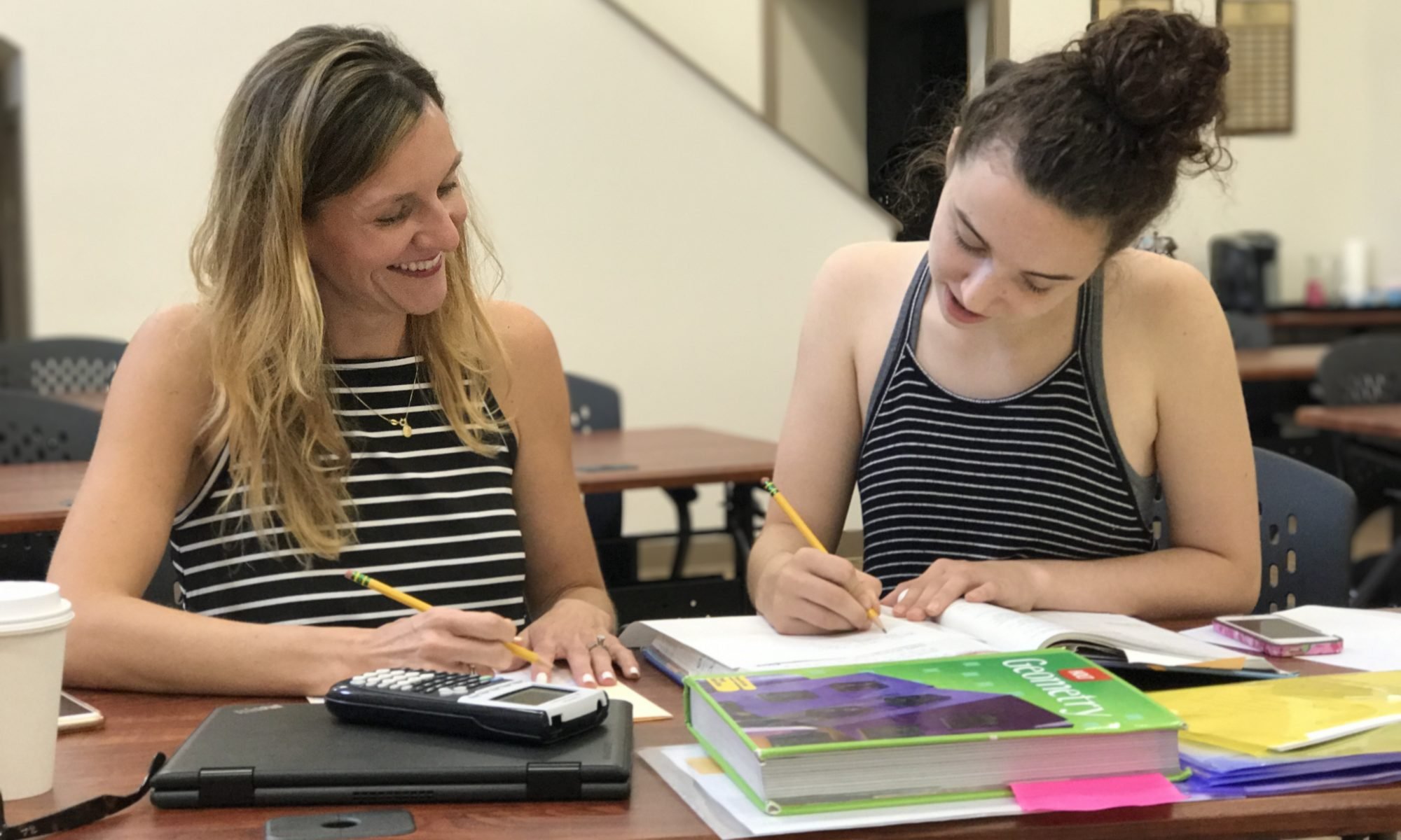 Two women sitting at a table studying together, smiling and writing in notebooks, surrounded by textbooks, notebooks, and a cup of coffee.