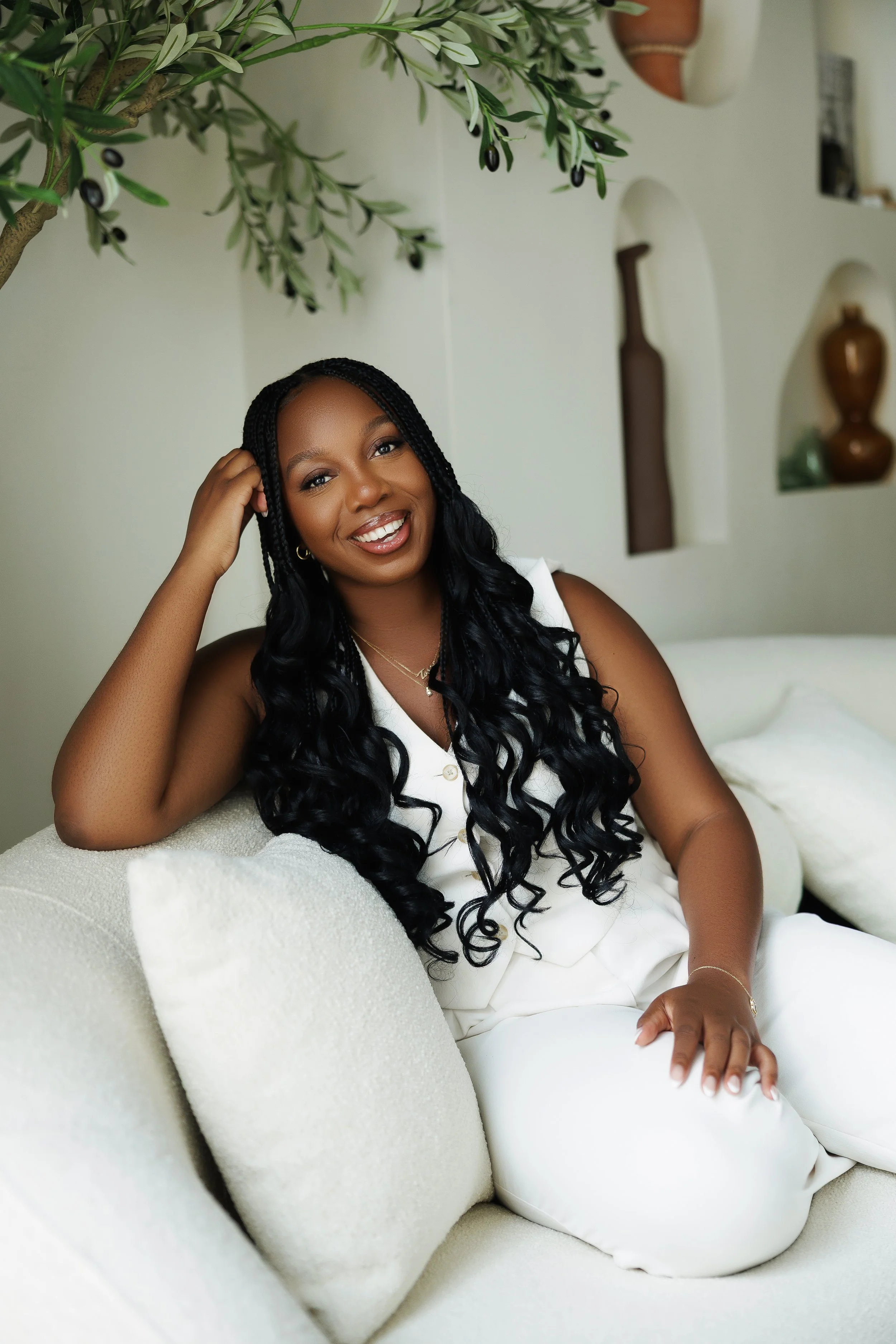 A woman with dark curly braided hair, wearing gold jewelry and a white outfit, sitting on a white sofa in a bright room with decorative shelves and plants.