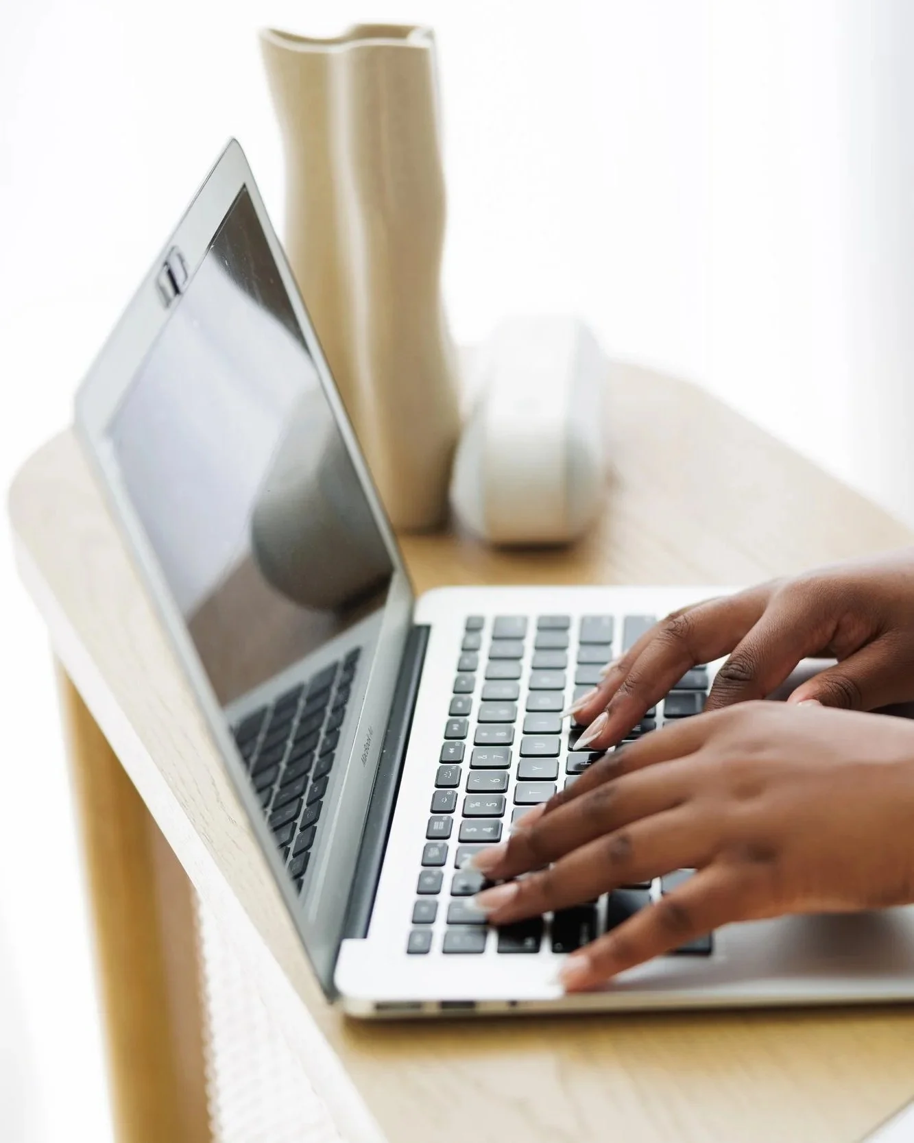 Person typing on a silver laptop with a wooden table, a white mug, a beige vase, and a small white speaker in the background.