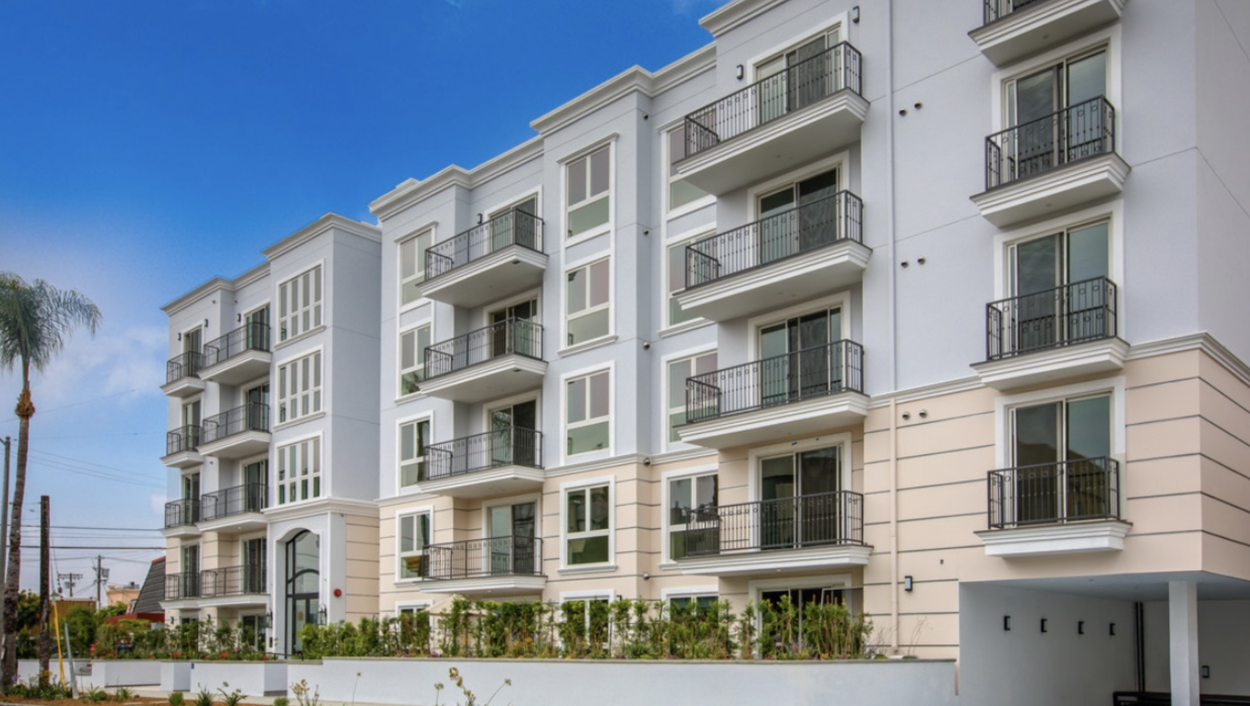 Modern white apartment building with multiple balconies and large windows, set against a bright blue sky with a palm tree nearby.