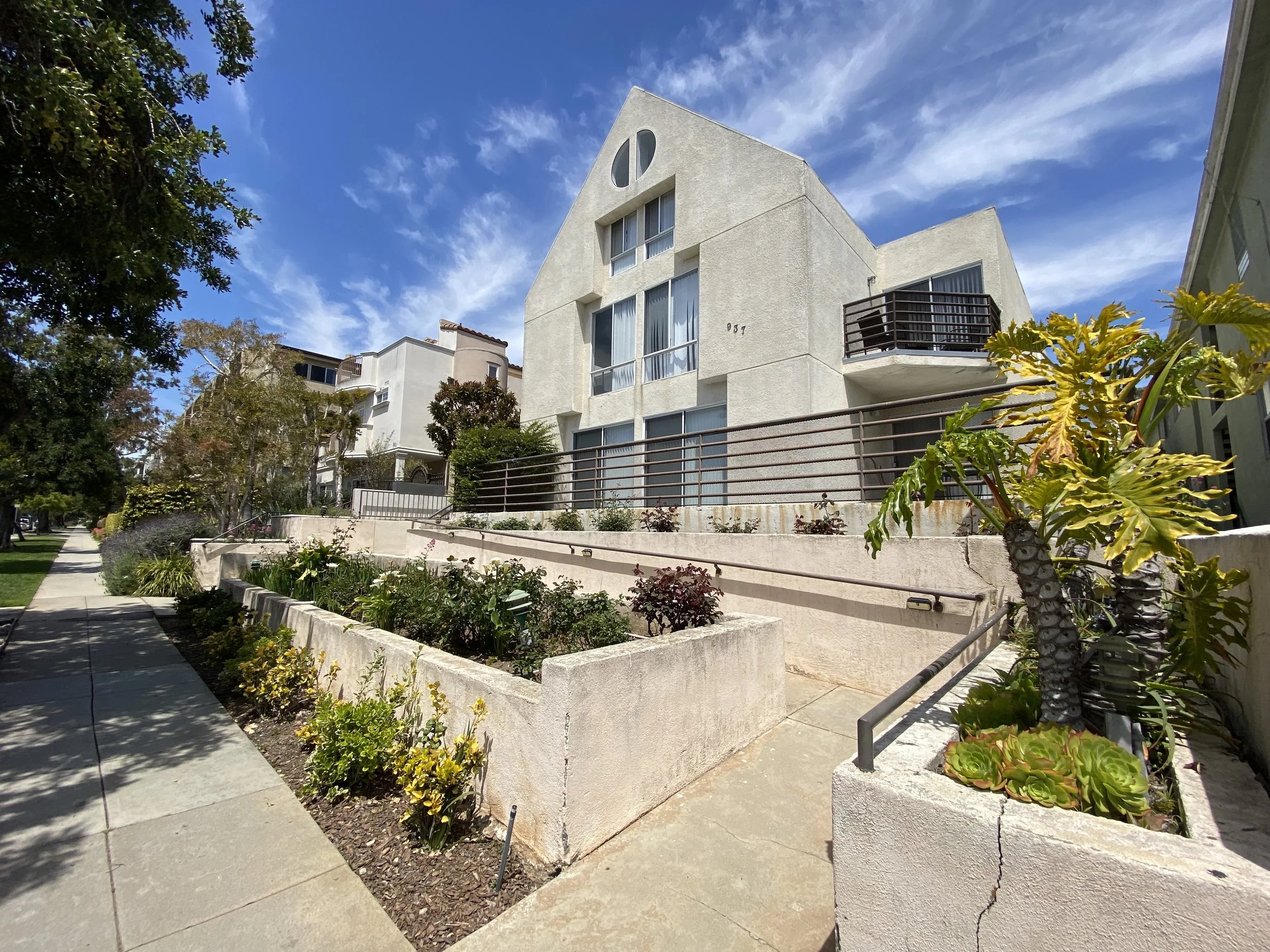 White modern residential building with balconies, set against a bright blue sky with clouds, adjacent to a sidewalk with a landscaped garden and trees.