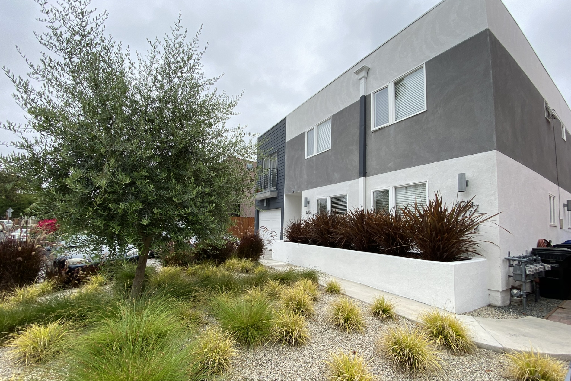 A modern multi-story residential building with white and gray exterior walls, multiple windows with blinds, and a small balcony on the second floor. In front, there is landscaped garden with various drought-tolerant plants, including a large bush, or