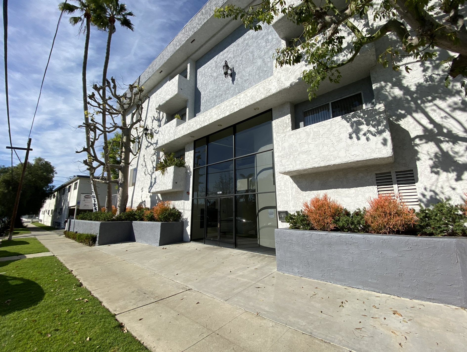 Modern white apartment building with large glass windows and balconies surrounded by trees and shrubs, under a partly cloudy sky.