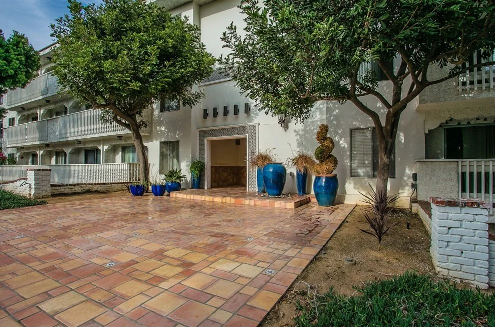 Apartment building entrance with a large tiled patio, potted plants, and trees.