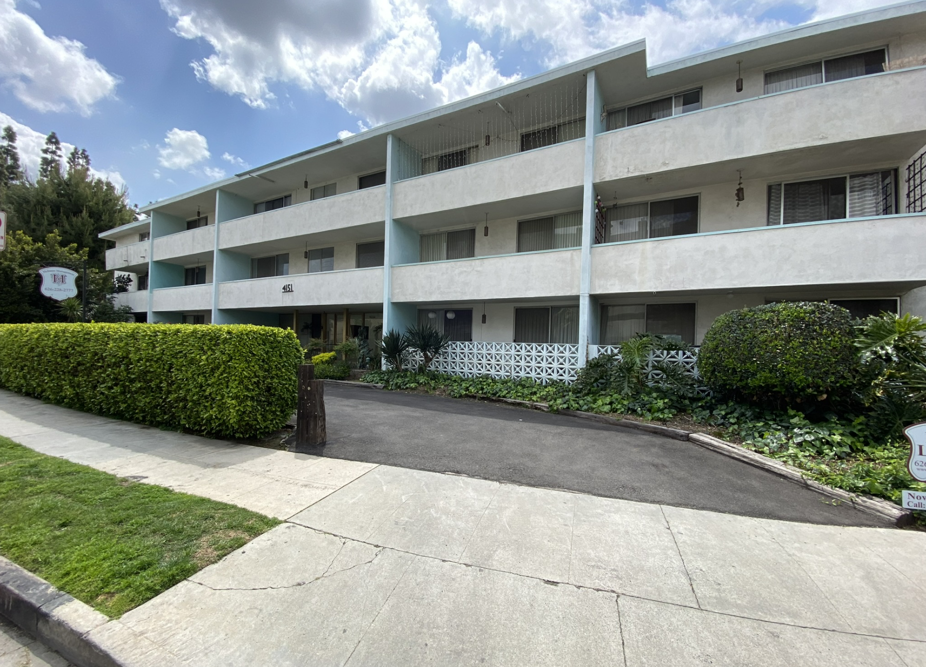 A three-story apartment building with a light-colored exterior, balconies with glass railings, and hanging lights. There are shrubs and plants in front, a sidewalk, and a cloudy sky overhead.
