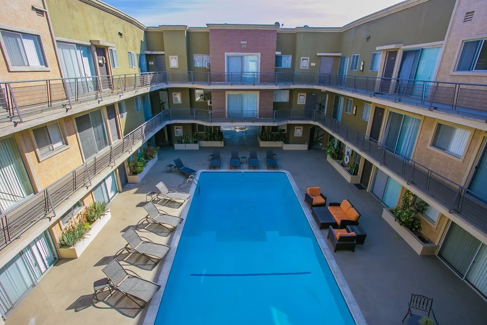 Indoor motel courtyard with a rectangular swimming pool, surrounded by lounge chairs, tables, and patio furniture, with warehouse-style apartment rooms on three sides and a clear sky overhead.