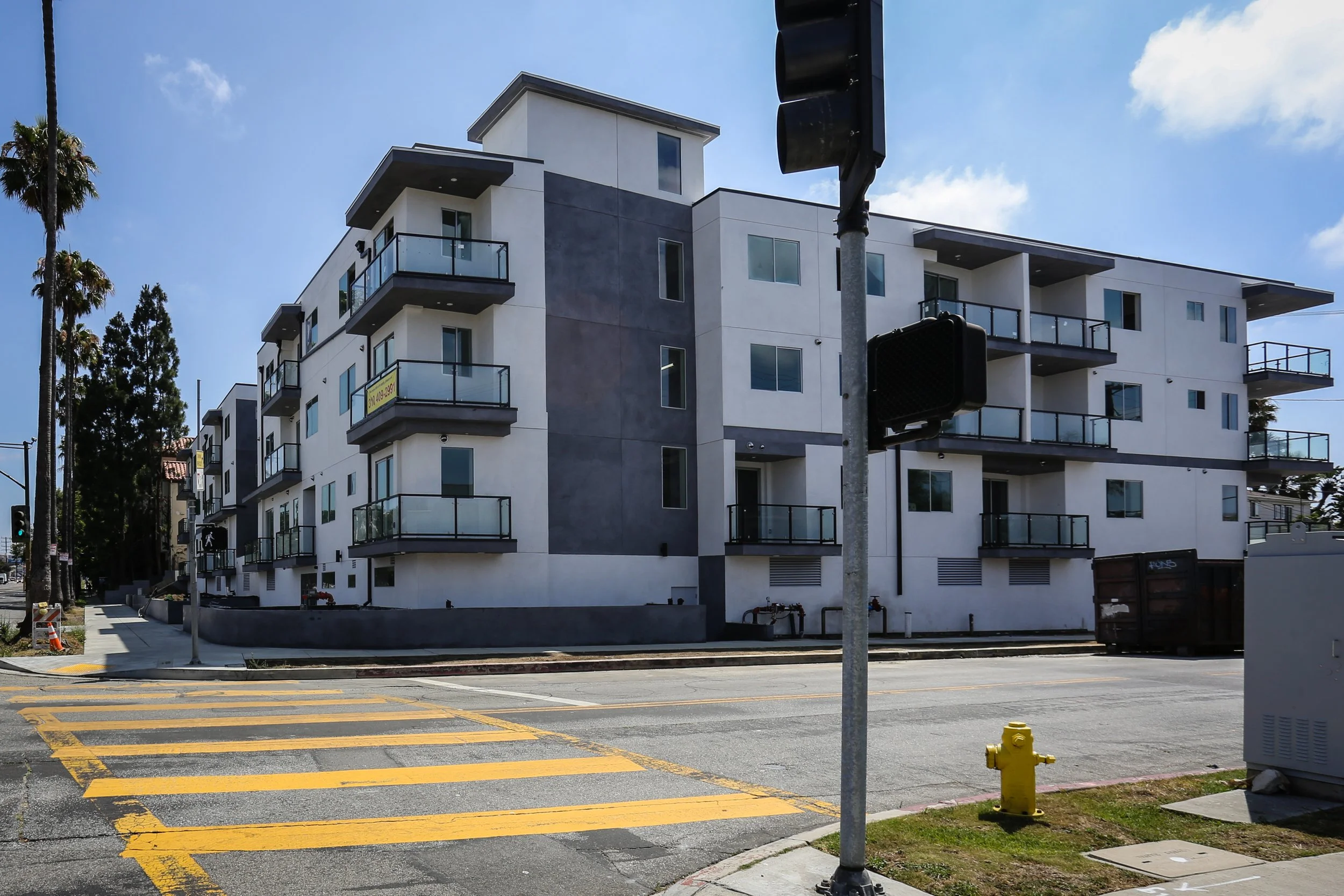 Modern white apartment building with multiple balconies, trees, and a blue sky, seen from the pedestrian crosswalk.