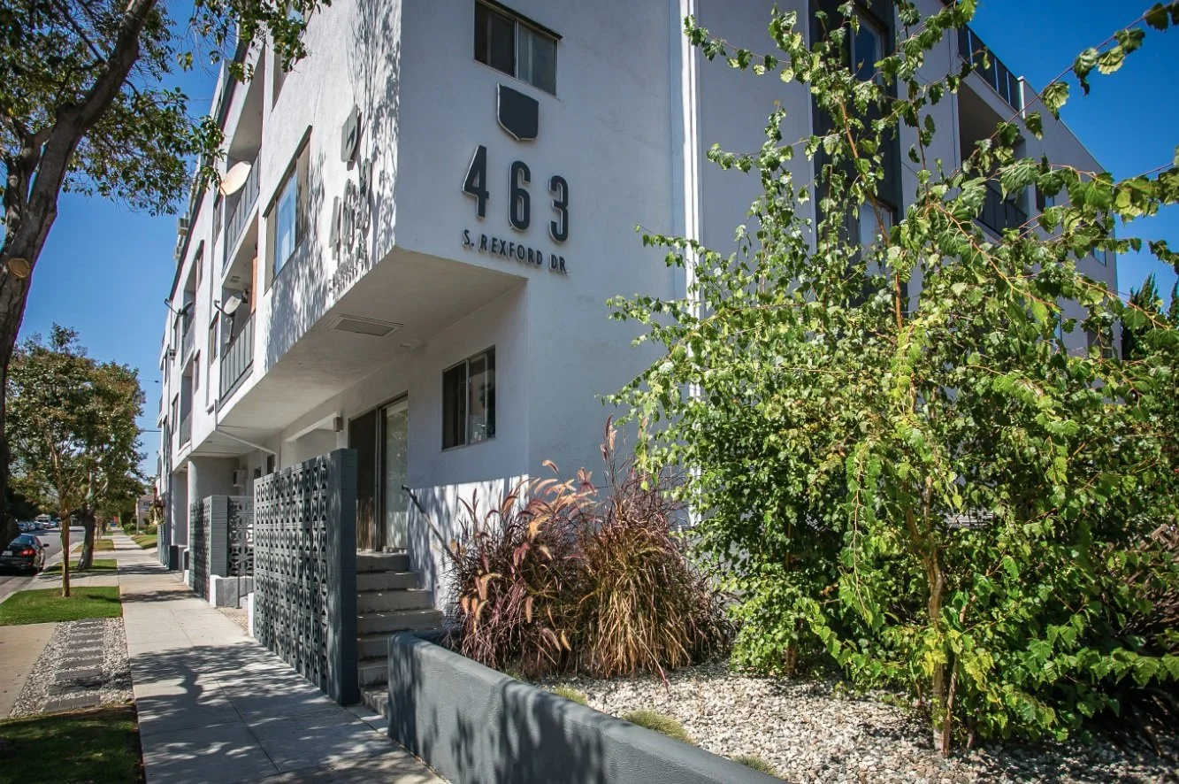 Exterior of a white apartment building with the address 463 S. Rexford Dr displayed on the wall, surrounded by trees and plants, under a clear blue sky.