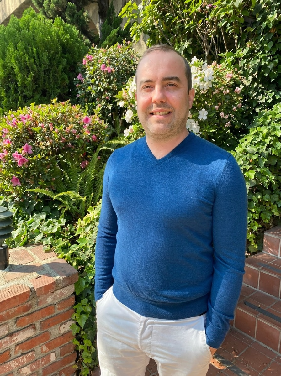 A man stands outdoors in front of lush green bushes and flowers, wearing a blue sweater and white pants, smiling.