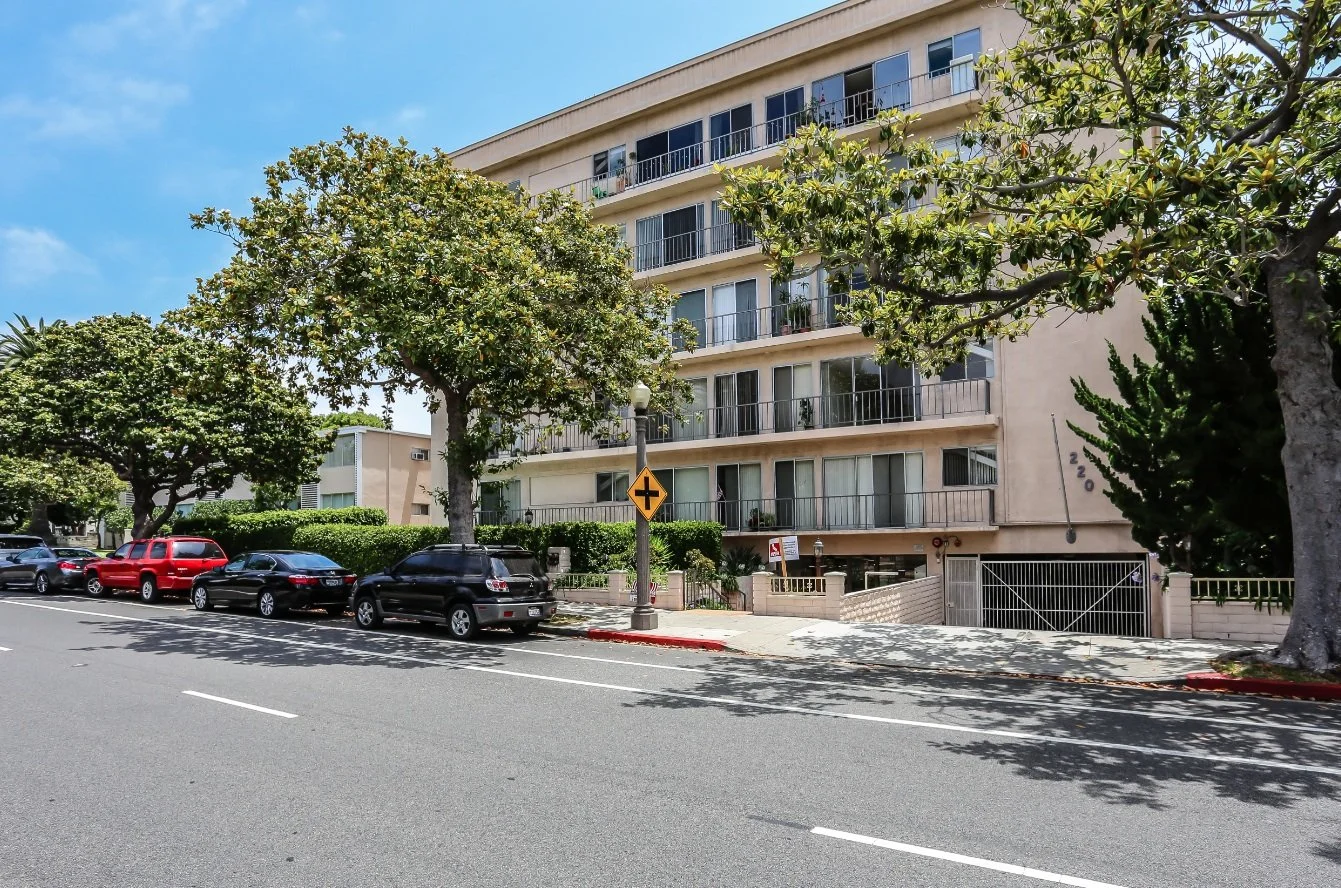 Street view of a beige apartment building with multiple balconies, parking lot with parked cars, trees lining the sidewalk, and a yellow street sign.