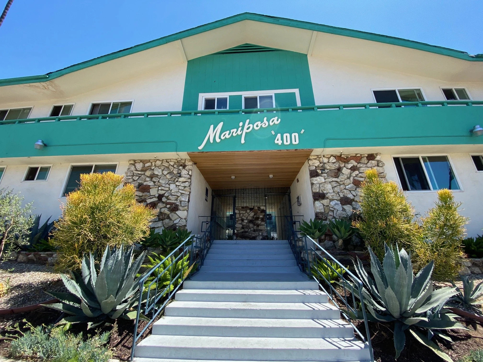 Exterior view of a building named Mariposa 400 with a stone facade, green accents, and a staircase leading up to the entrance, surrounded by desert plants and greenery.
