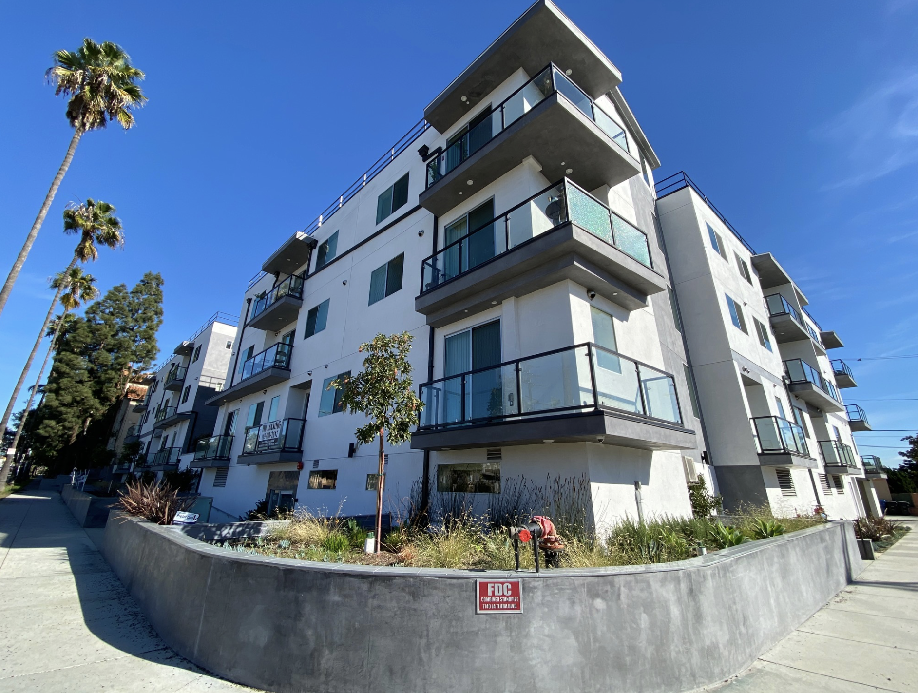A modern white apartment building with multiple balconies and glass railings, surrounded by palm trees and green landscaping, under a clear blue sky.
