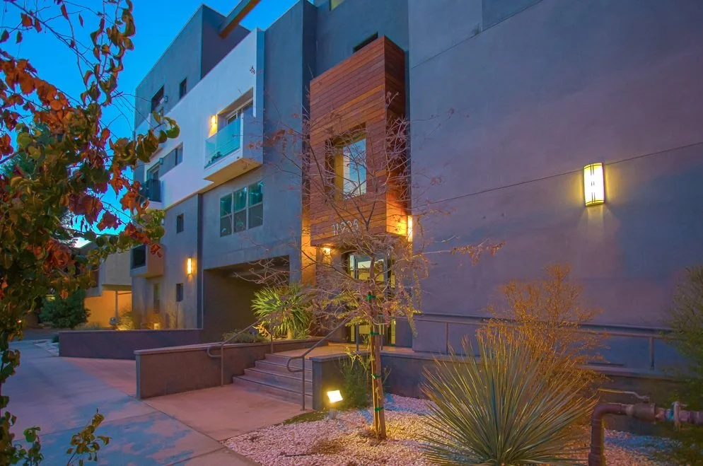 Exterior view of a modern apartment building during dusk, with warm exterior lighting, stairs, and landscaping including trees and desert plants.