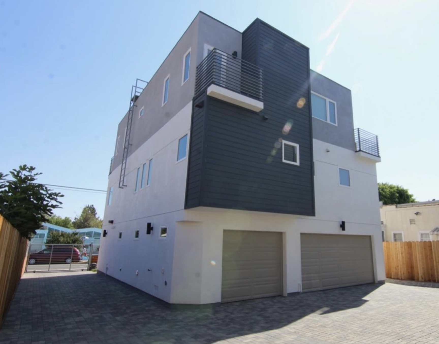 Modern multi-story residential building with a white and dark gray exterior, featuring balconies, multiple windows, and a garage with two doors. The sky is clear and sunny.