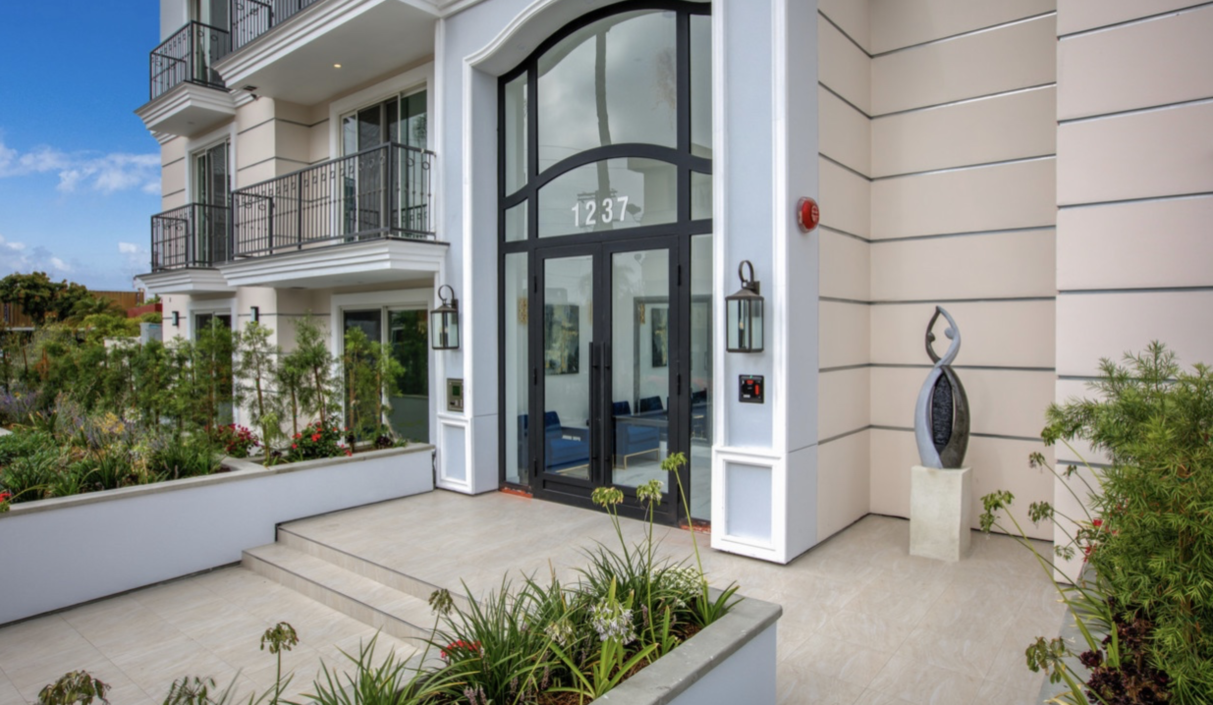 Modern multi-story apartment building with balconies, large glass entrance door with the number 1237, landscaped garden with plants and flowers, outdoor sculpture, and clear blue sky.