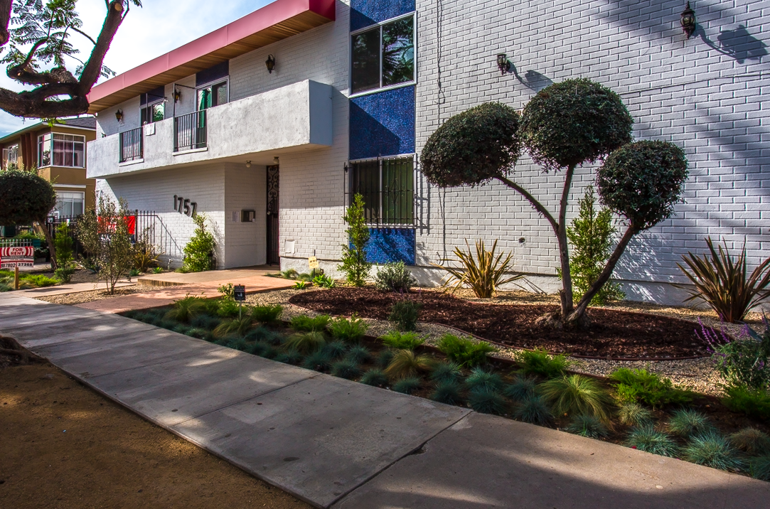 The exterior of a white brick apartment building with a red roof trim, featuring a small garden with trimmed bushes and plants, a concrete walkway, and a visible address number 1757.