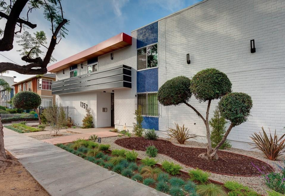 Modern residential building with white brick exterior, blue accents, and a red overhang. Front yard landscaped with bushes, trees, and ground cover, with a sidewalk in the foreground.