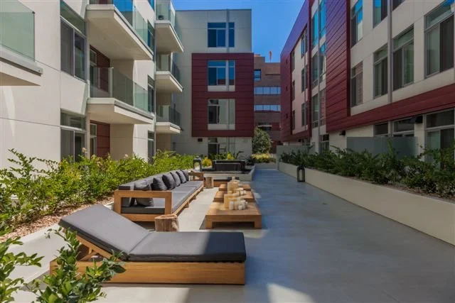 Modern apartment courtyard with outdoor seating, low wooden tables, and green plants surrounding the area.