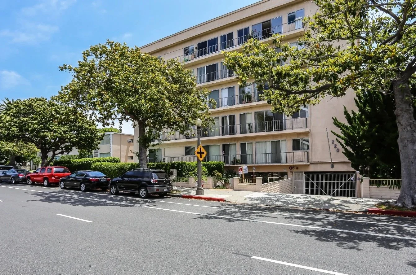 Street view featuring parked cars, trees, a lamp post, and a beige apartment building with balconies.
