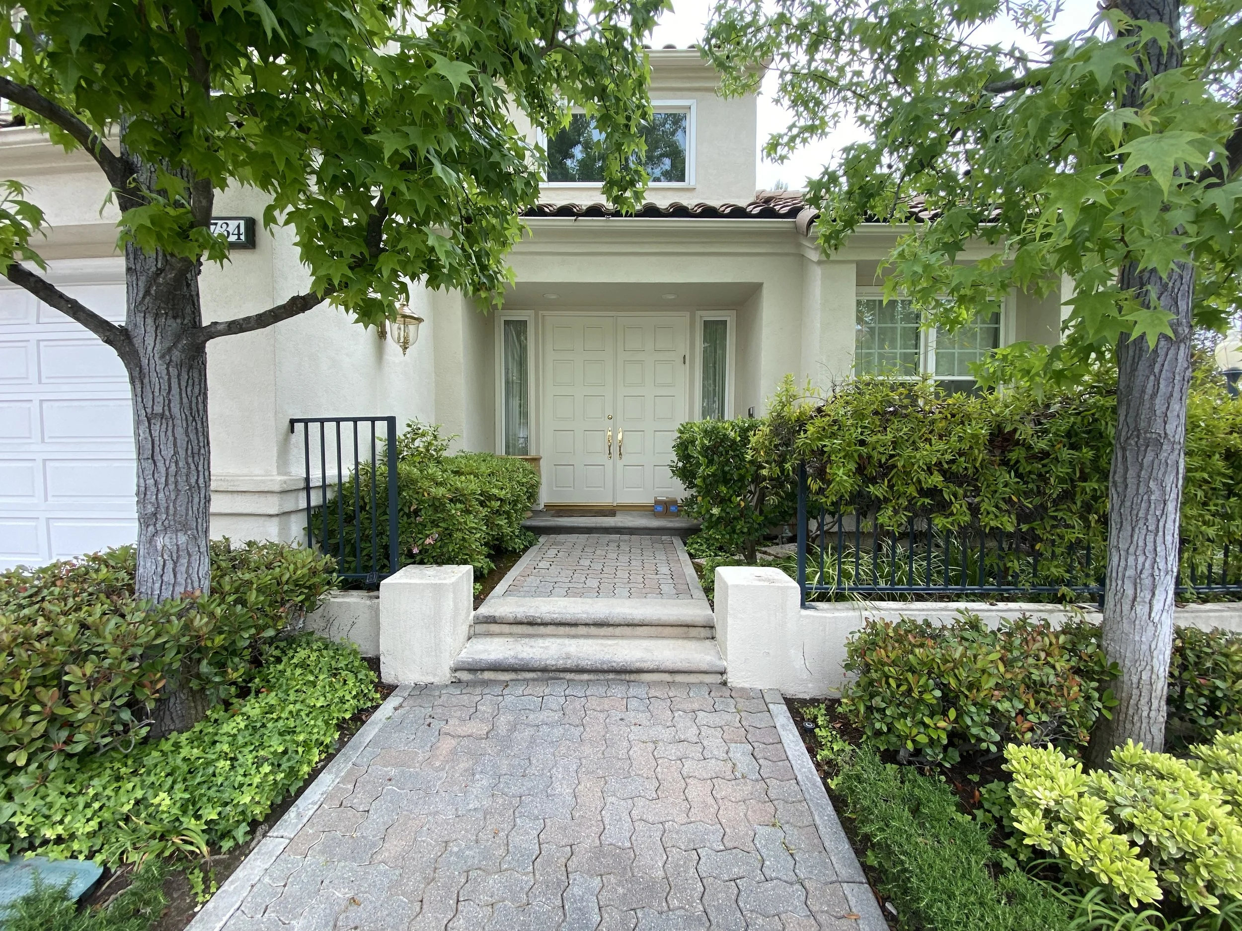 Front view of a house with two trimmed trees, a paved pathway, shrubs, and windows.