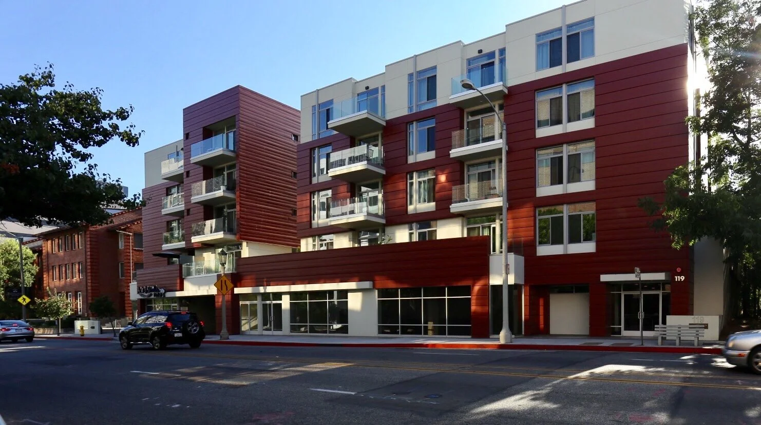 Modern multi-story residential building with red and white exterior, large windows, and balconies, adjacent to a street with cars and trees.